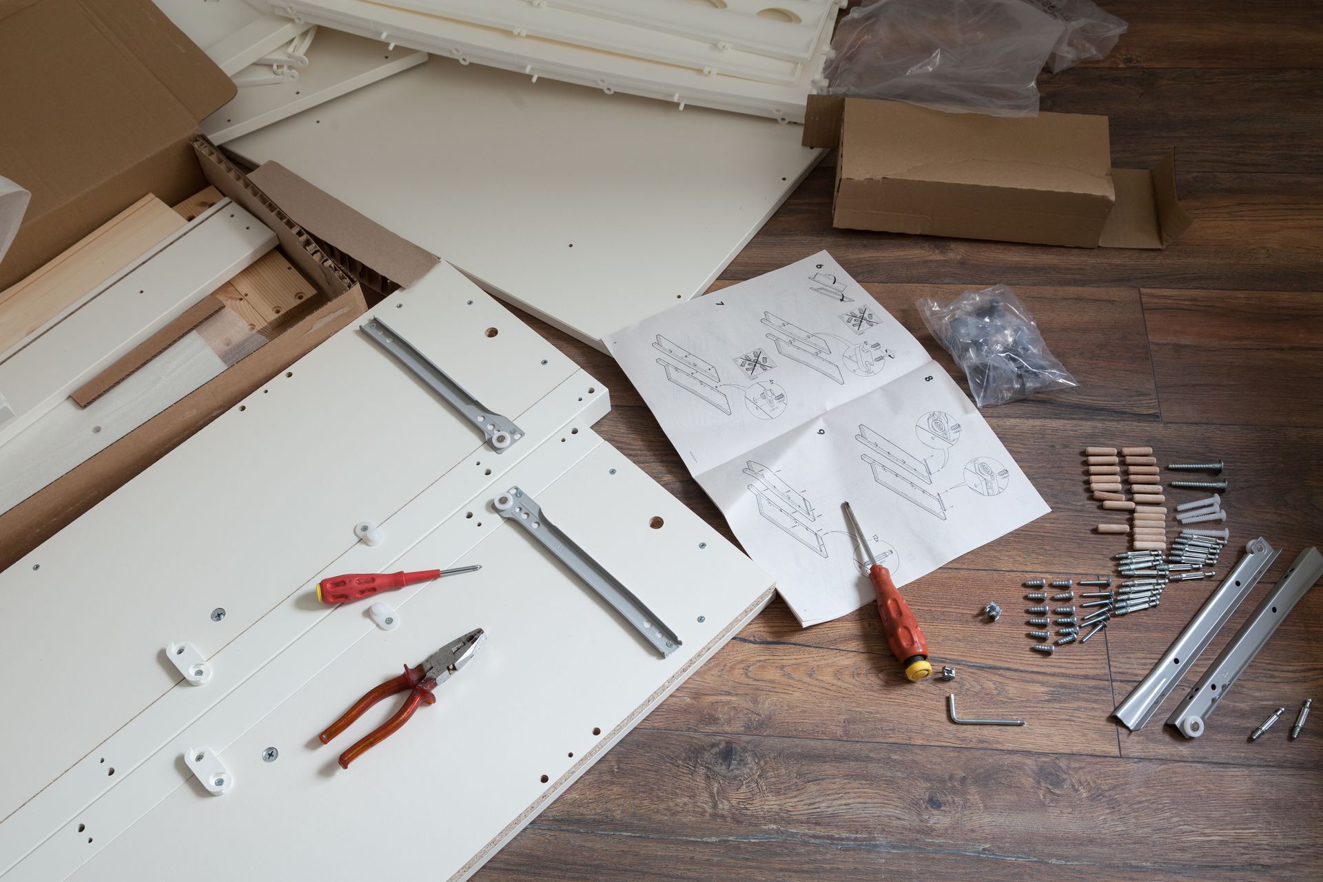 Furniture assembly in progress on a wood floor with white panels, tools, instruction sheets, and hardware scattered around.