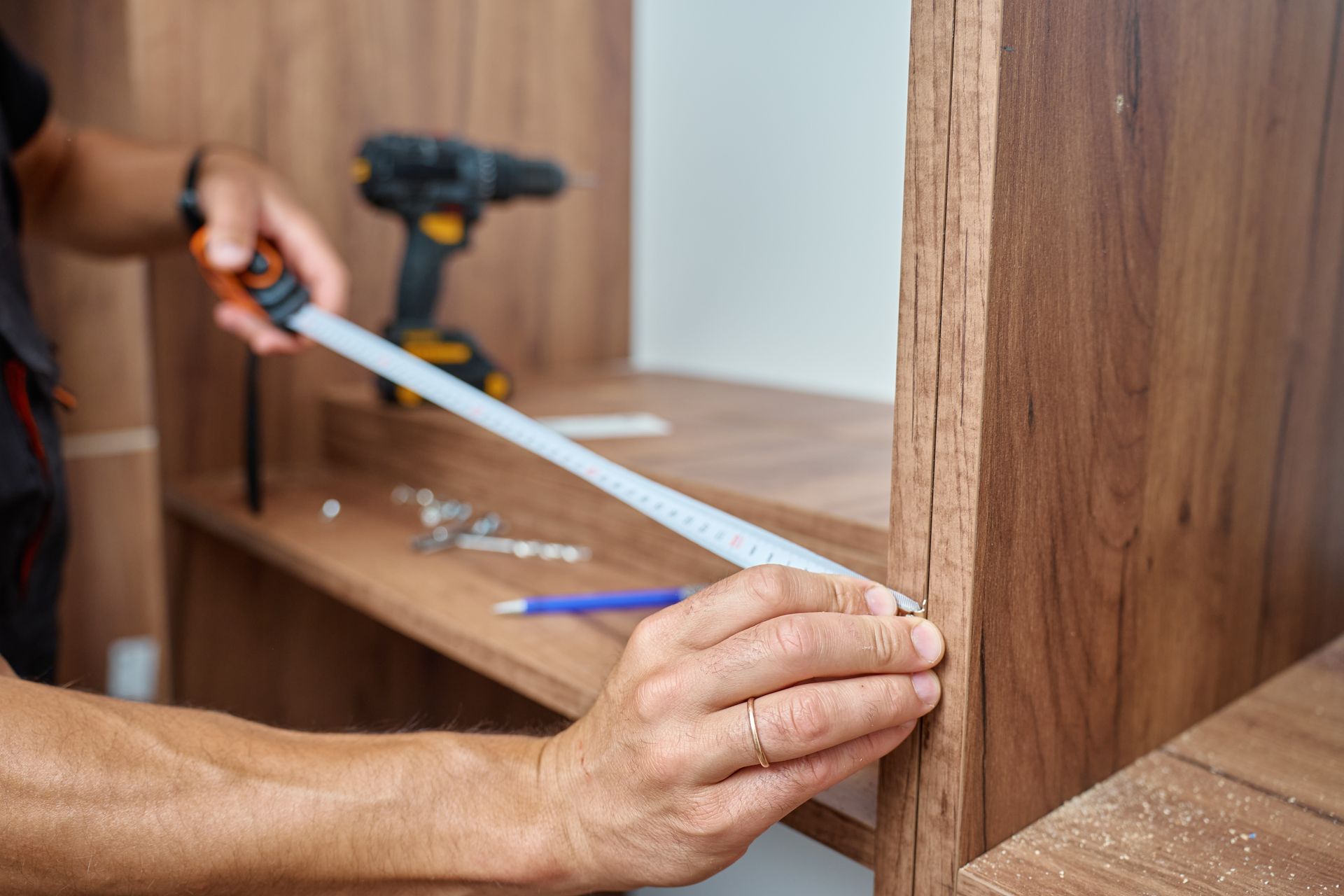A person measures a wooden shelf with a tape measure, with a cordless drill and tools nearby on a workbench.