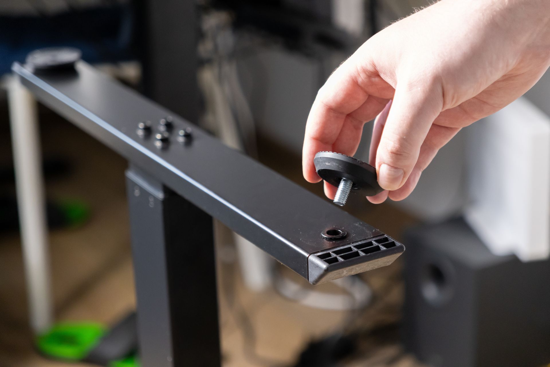 A hand holds a leveling foot above a mounting hole on the metal leg of a desk frame.