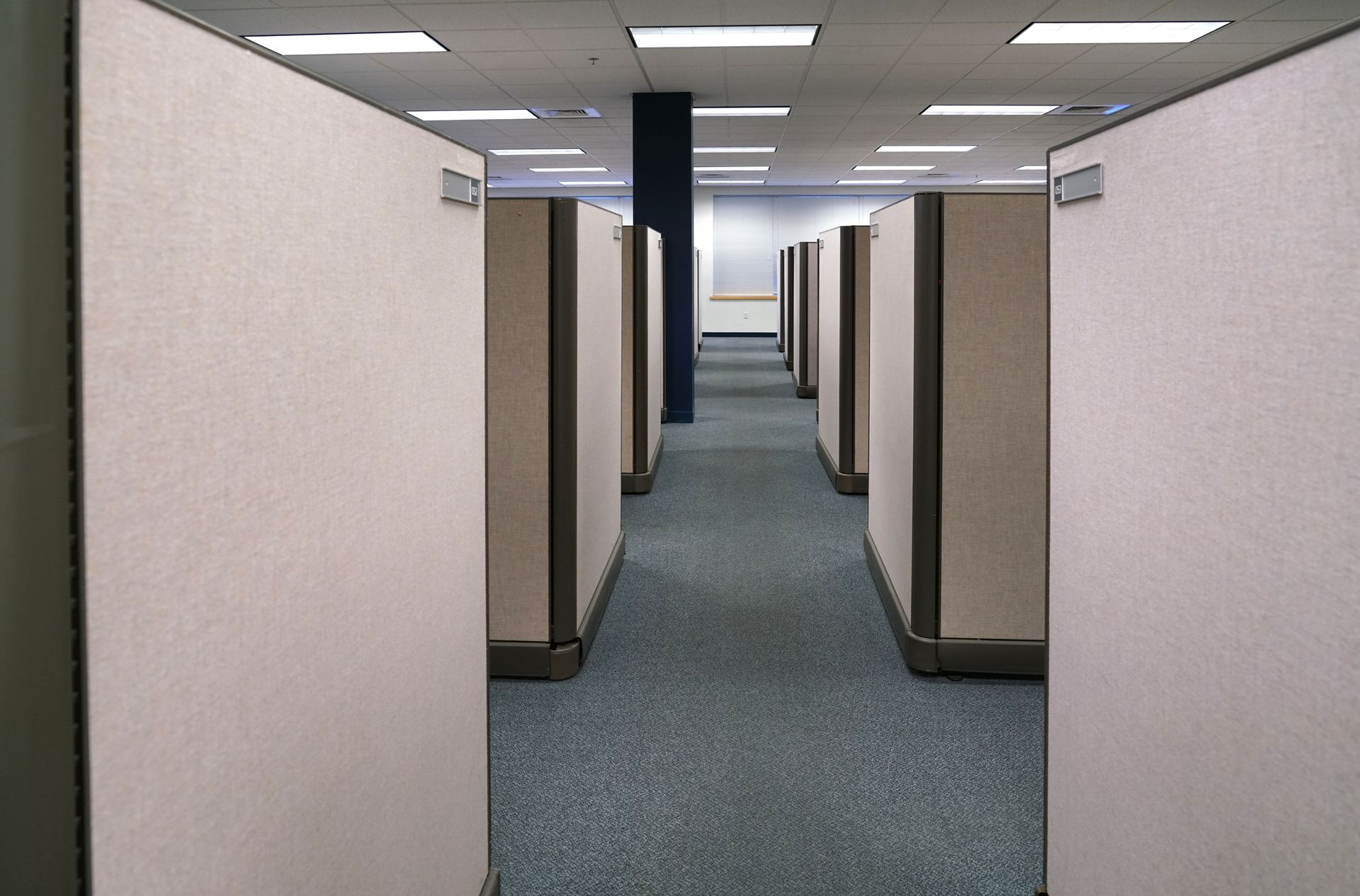 An aisle between beige office cubicle walls leading to a dark pillar in a brightly lit, carpeted office space.