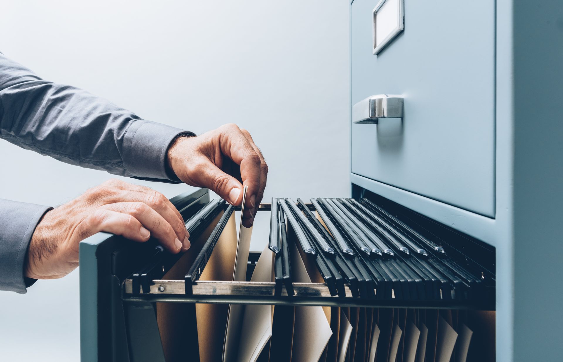 A person in a gray shirt searching through hanging file folders inside an open metal filing cabinet drawer.