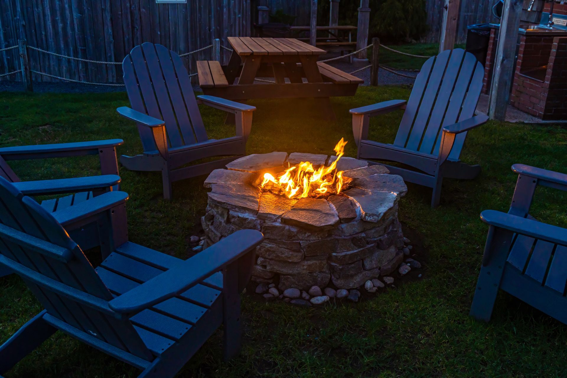 A stone fire pit with a glowing fire surrounded by dark wooden Adirondack chairs on a grassy lawn at dusk.