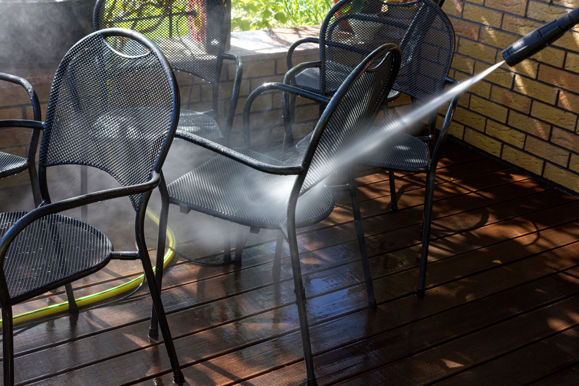 A pressure washer sprays water onto black metal patio chairs on a wooden deck.
