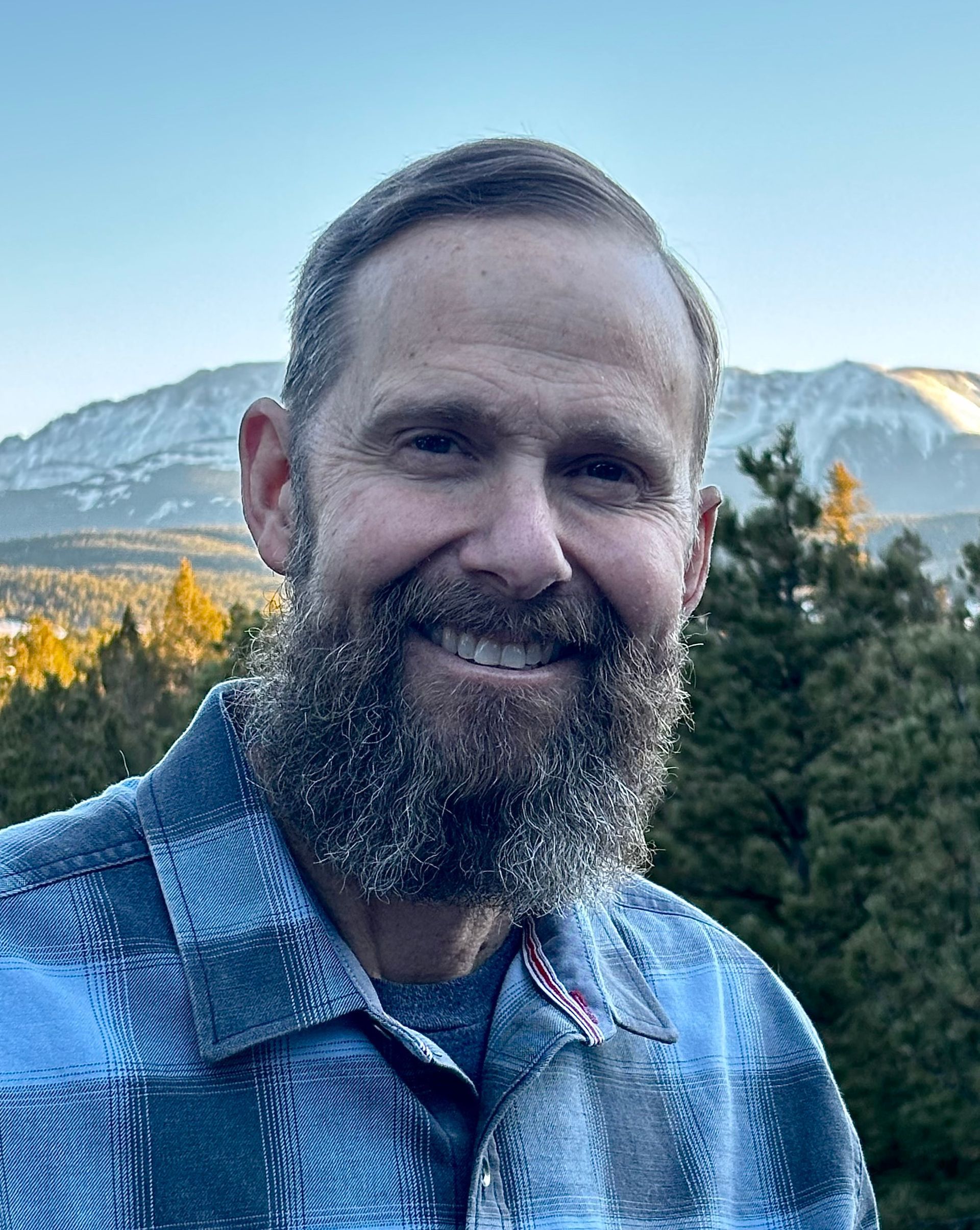 Man with beard smiles outside, wearing blue plaid shirt, mountains in background.