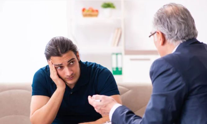 A man with a concerned expression consults with a therapist, indoors on a couch.