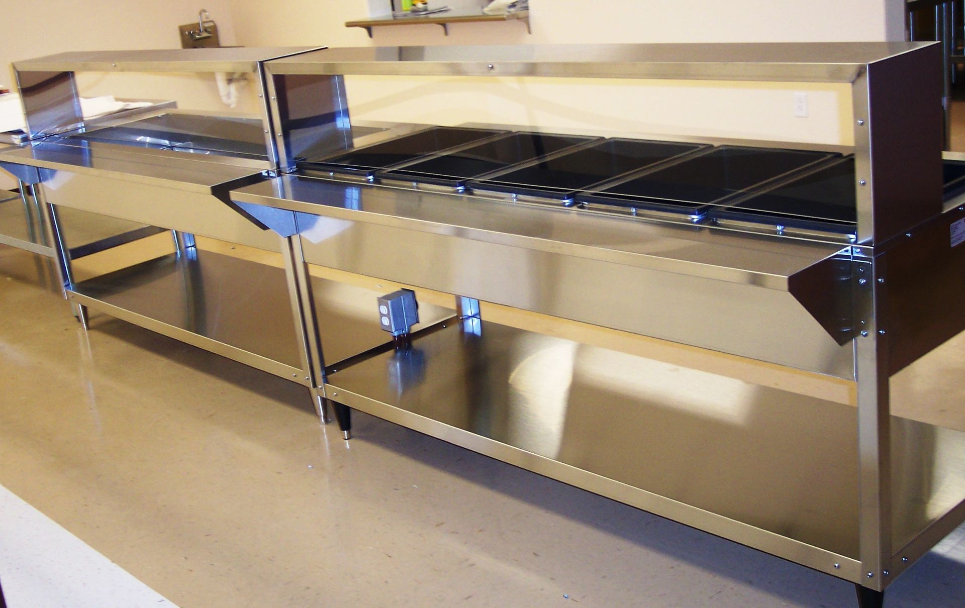 Two stainless steel commercial serving counters with overhead heat lamps and lower shelves in a kitchen setting.