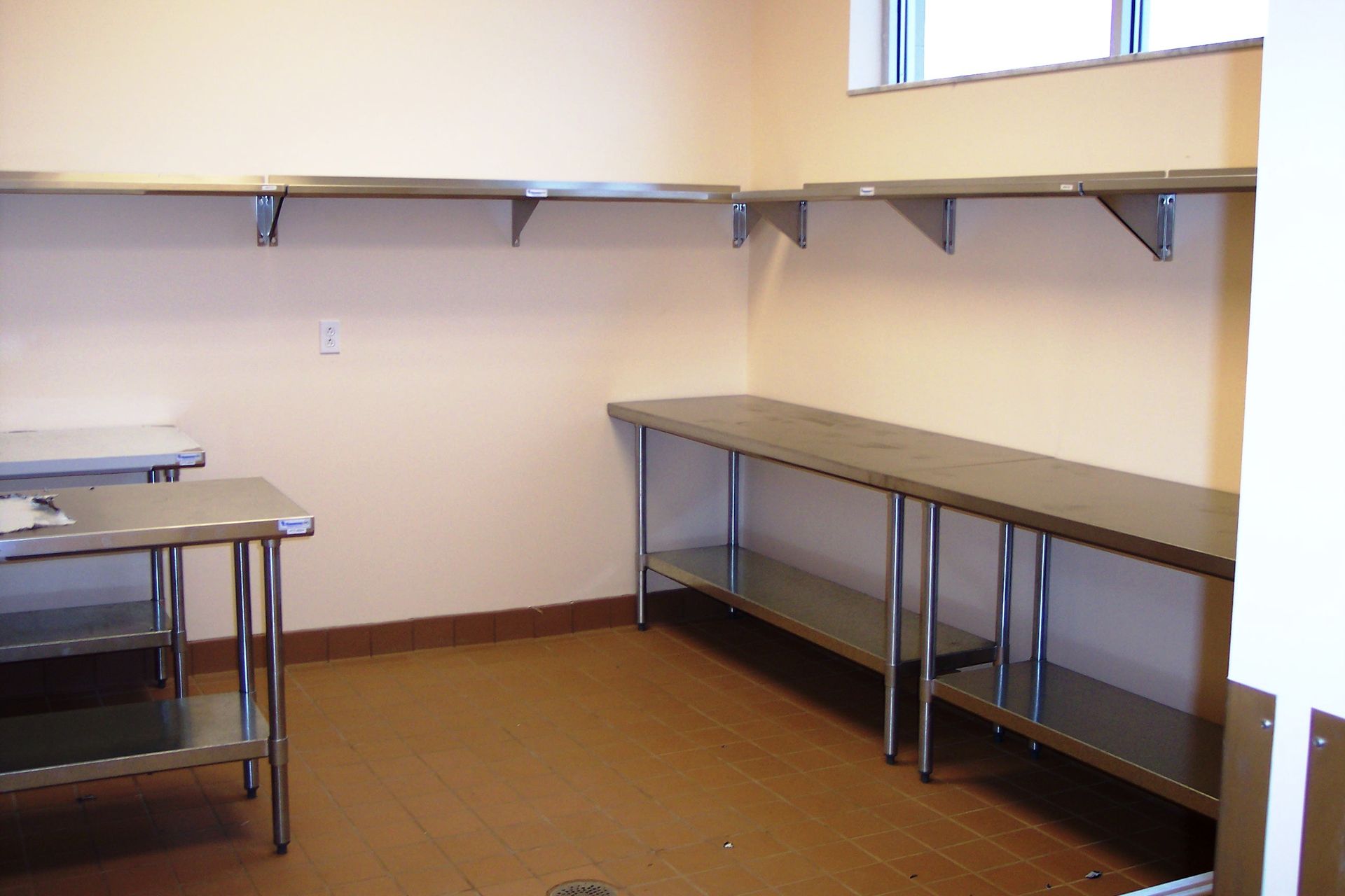 Stainless steel kitchen prep tables and wall-mounted shelving in an empty room with light-colored walls and tan flooring.