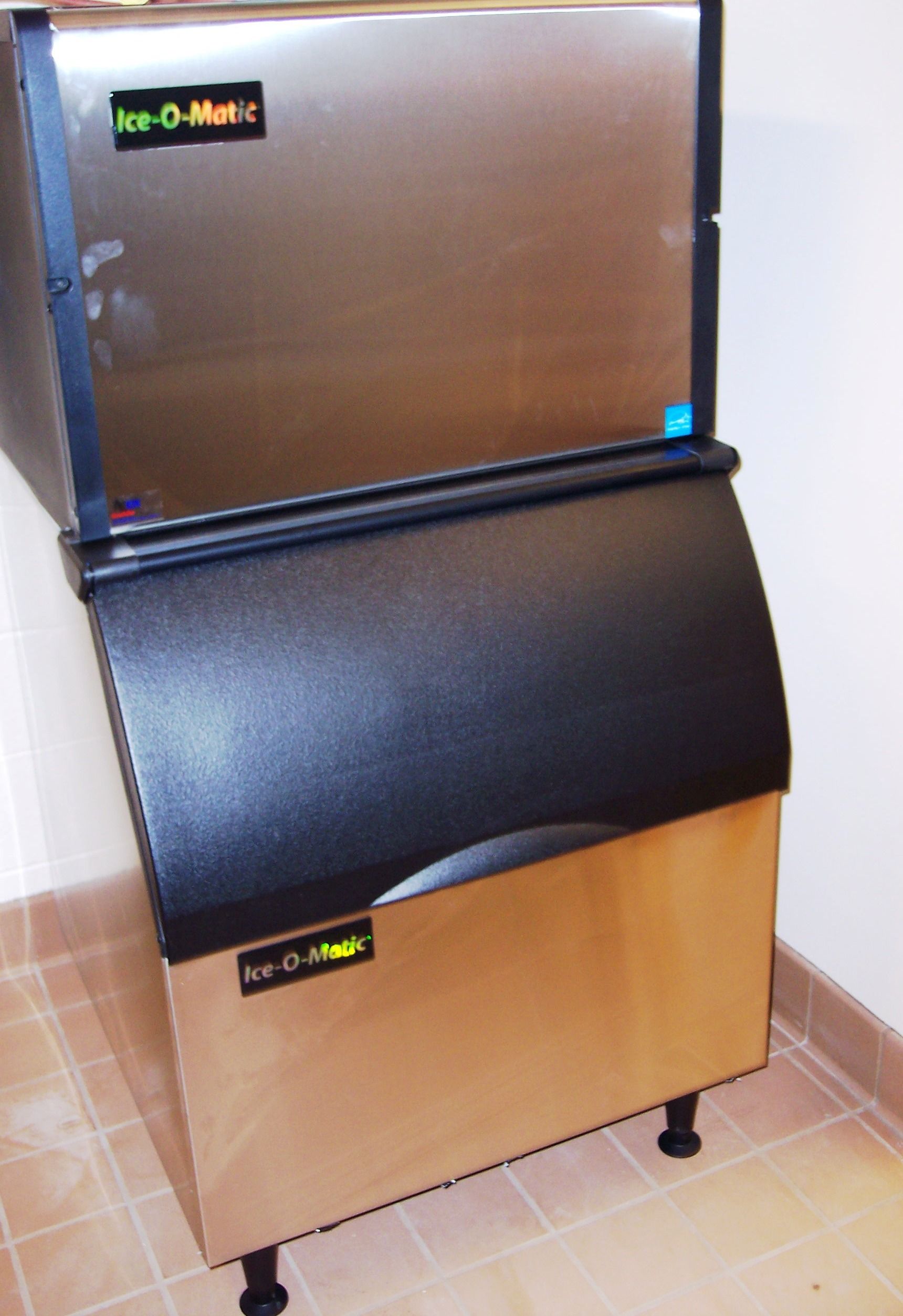 An Ice-O-Matic ice machine with a stainless steel top unit and a black bin with a golden-brown front panel.