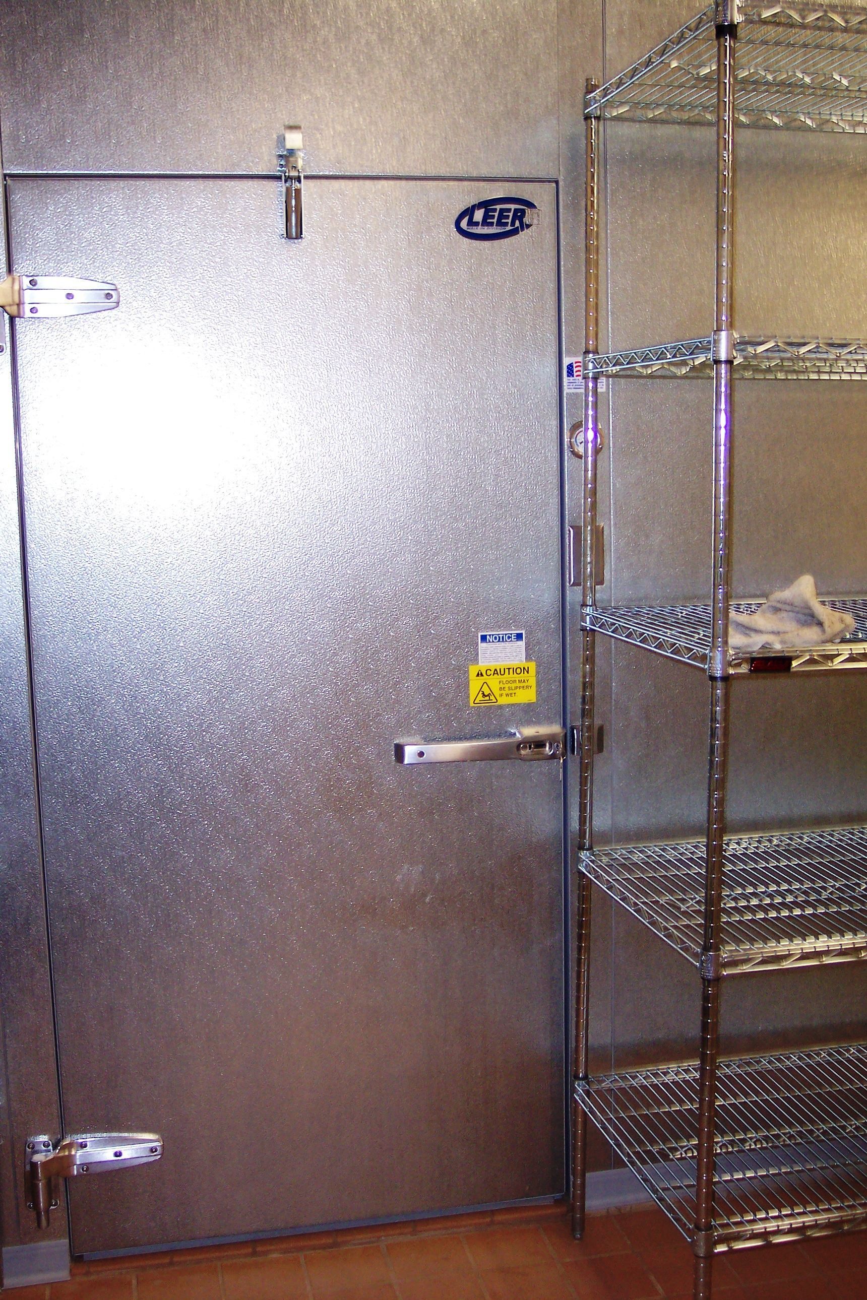 A stainless steel commercial walk-in freezer door with a heavy-duty latch, next to a metal wire shelving unit.