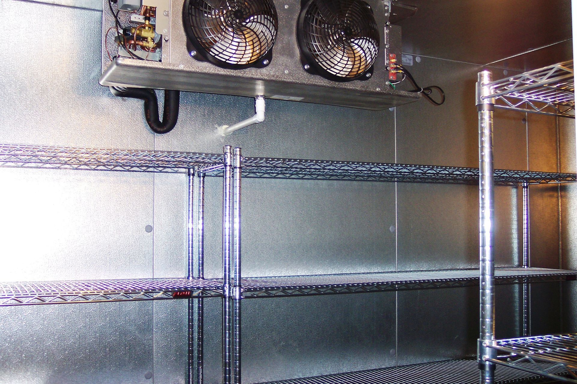 A commercial walk-in refrigerator interior with two fans mounted on the ceiling and metal wire shelving units.