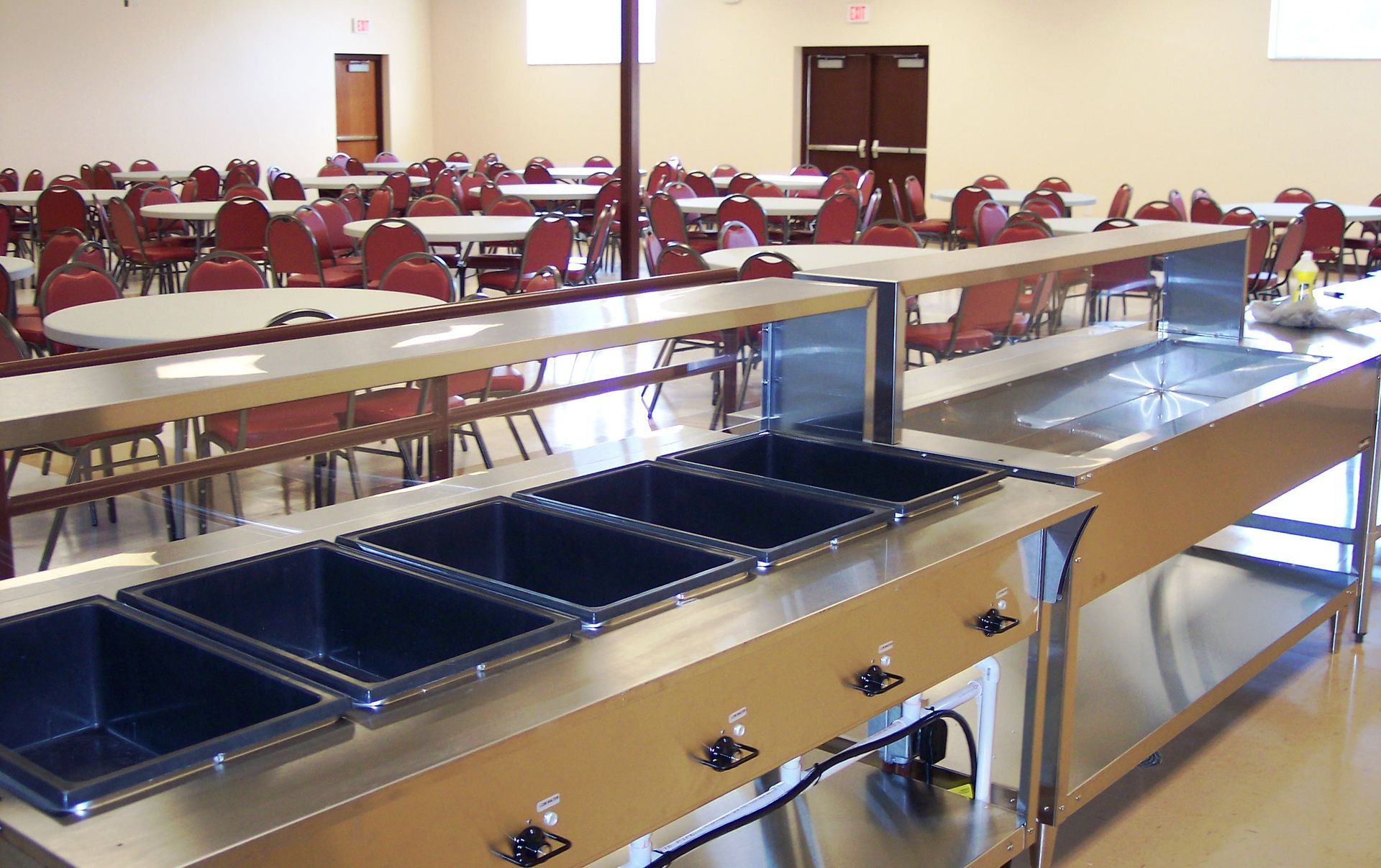 A stainless steel buffet serving station with four black food pans in a large, empty dining hall with tables and chairs.