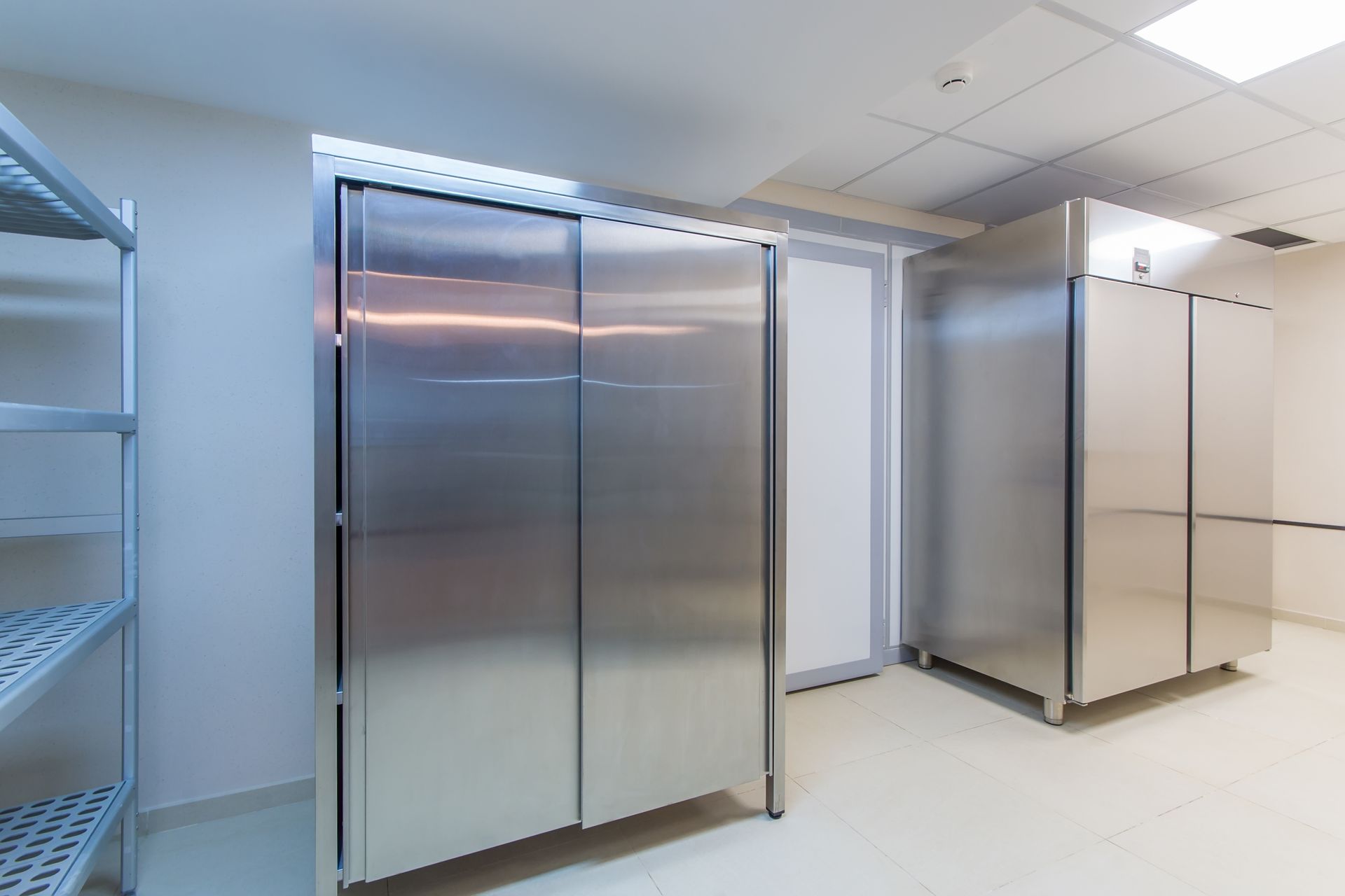 Two large, stainless-steel industrial refrigerators stand in a brightly lit, clean, white-walled commercial kitchen.