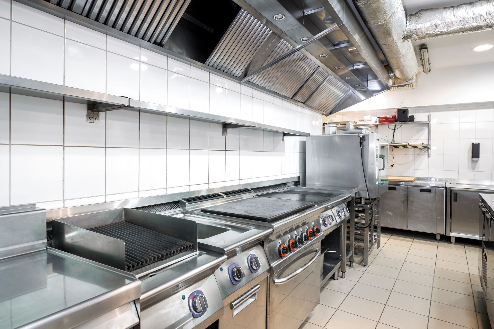 A clean, professional commercial kitchen with stainless steel appliances, a large exhaust hood, and white tiled walls.