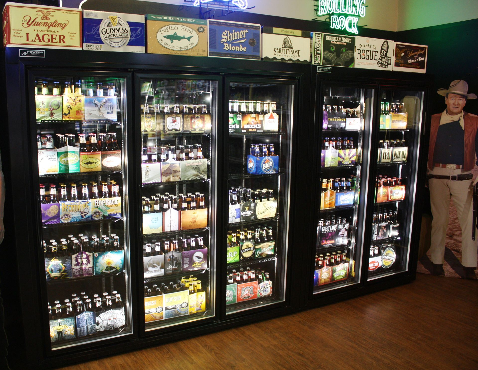 A wall of glass-door refrigerators filled with various beer bottles and six-packs, with a life-size cowboy cutout nearby.