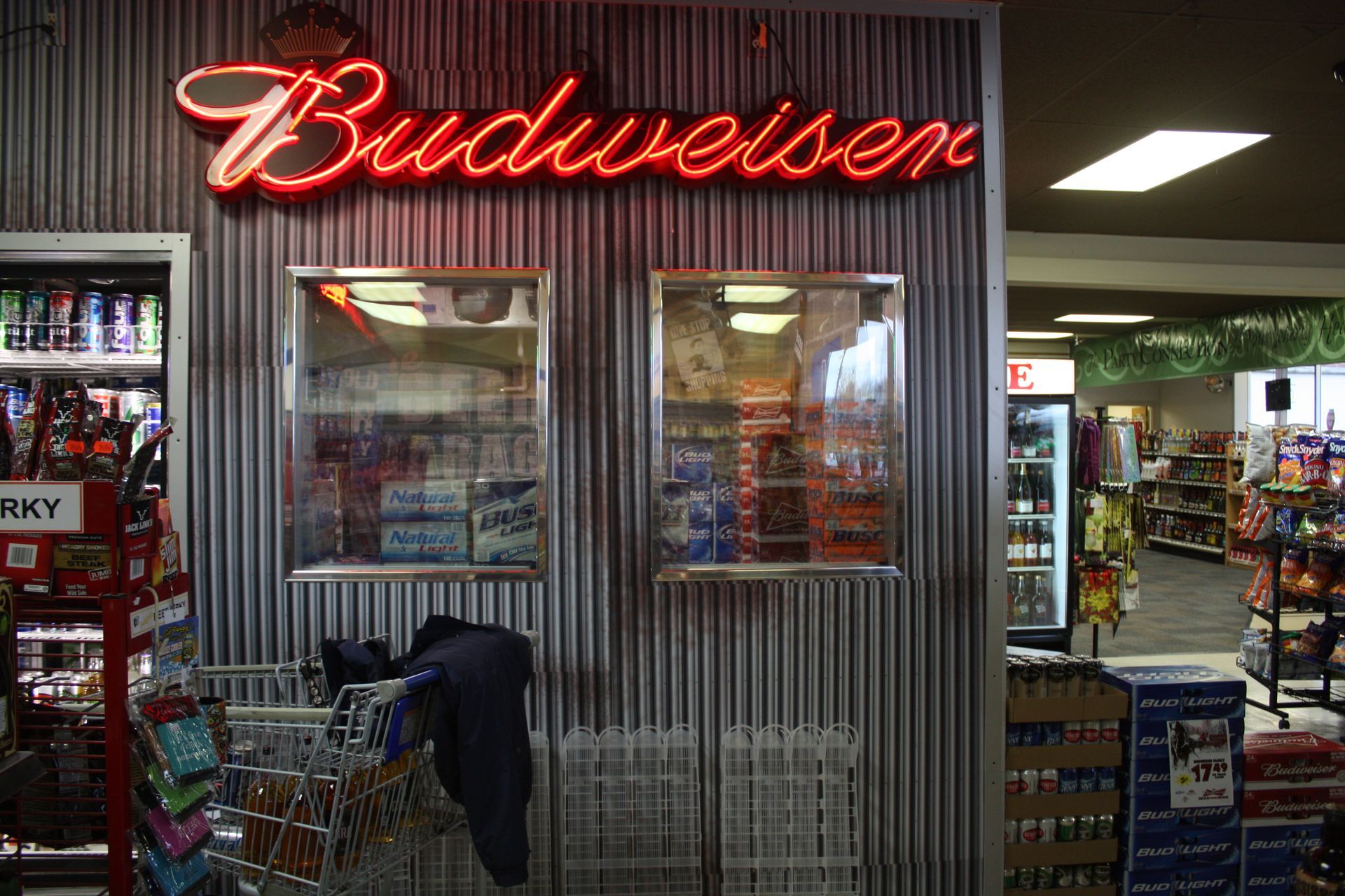 A Budweiser neon sign mounted on a gray corrugated metal wall above two display cases in a convenience store.