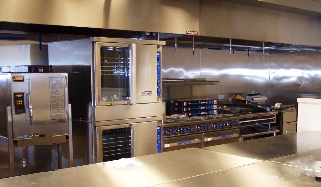 A professional stainless steel kitchen with commercial ovens, a stovetop range, and a large metal preparation counter.