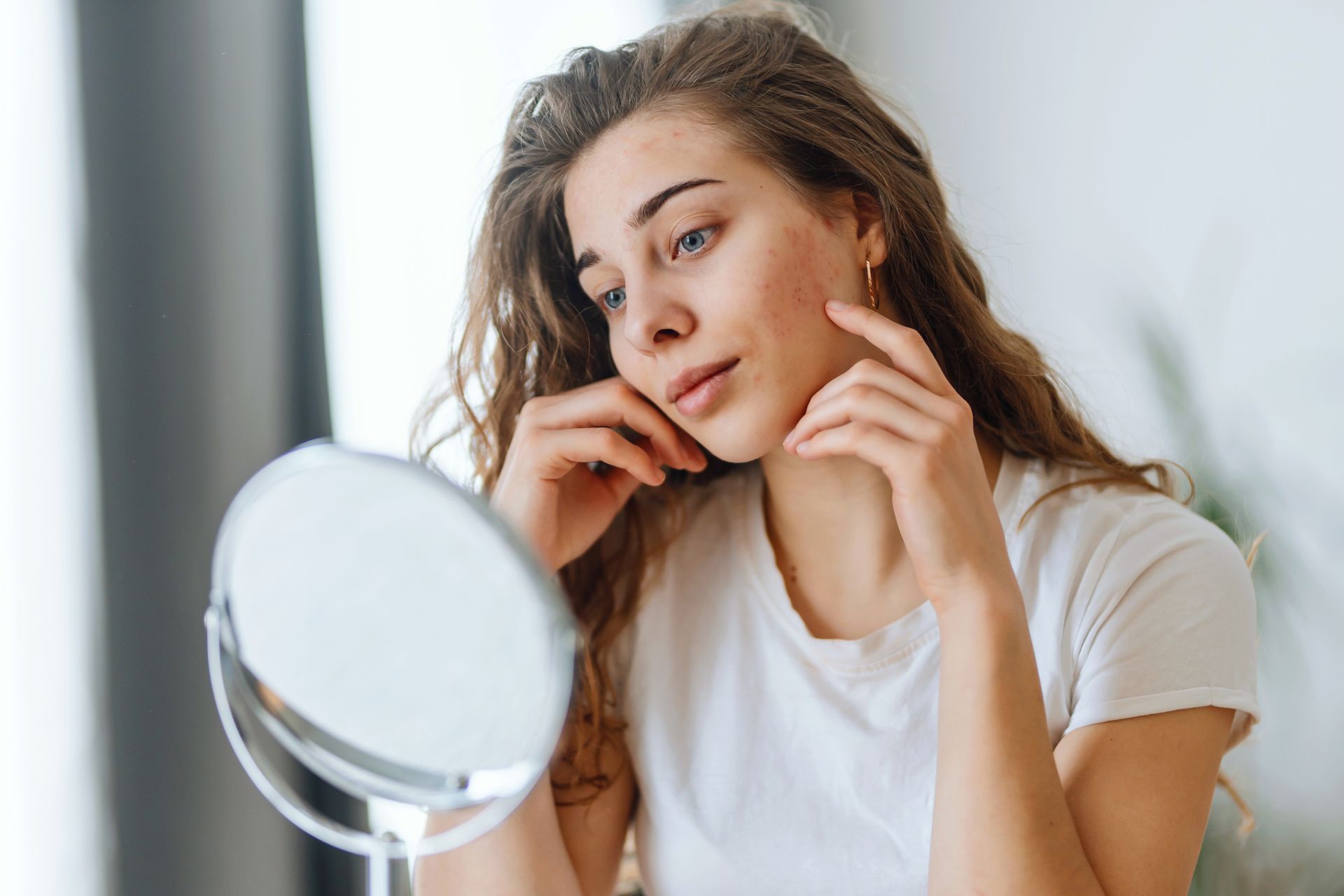 Woman with curly hair examines her face in a small mirror, touching her cheeks. Indoors, wearing a white shirt.