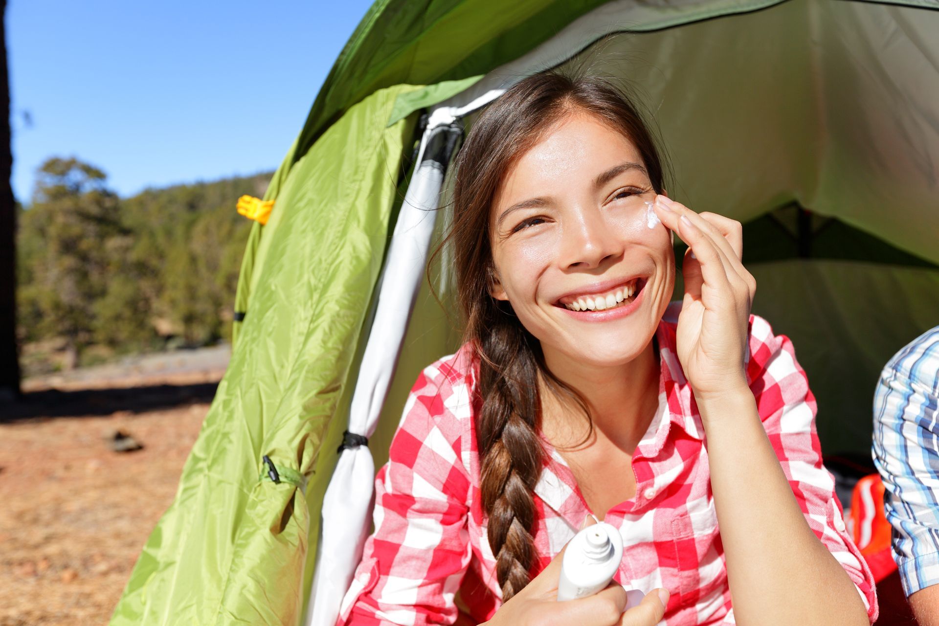 Woman in red plaid shirt smiling, applying sunscreen outside a tent.