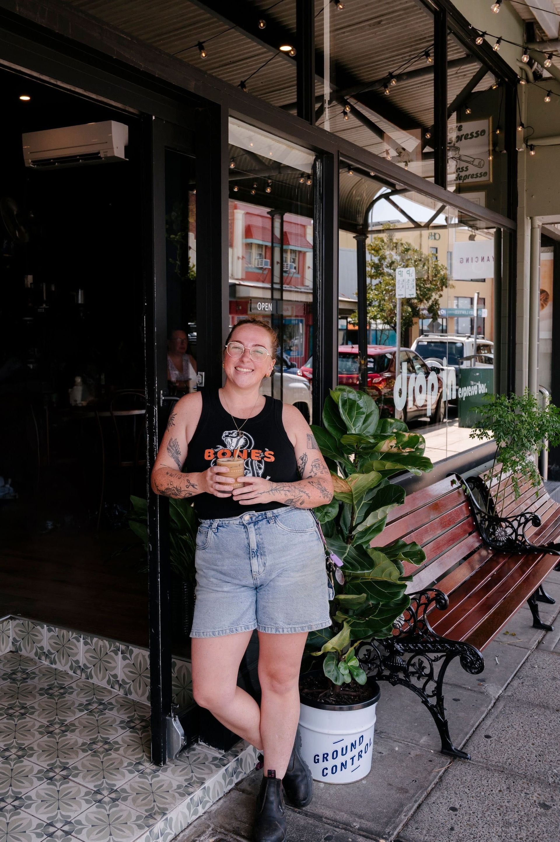 Woman in denim shorts and a tank top holding a drink, standing outside a cafe — Drop In Espresso Bar in Wallsend, NSW