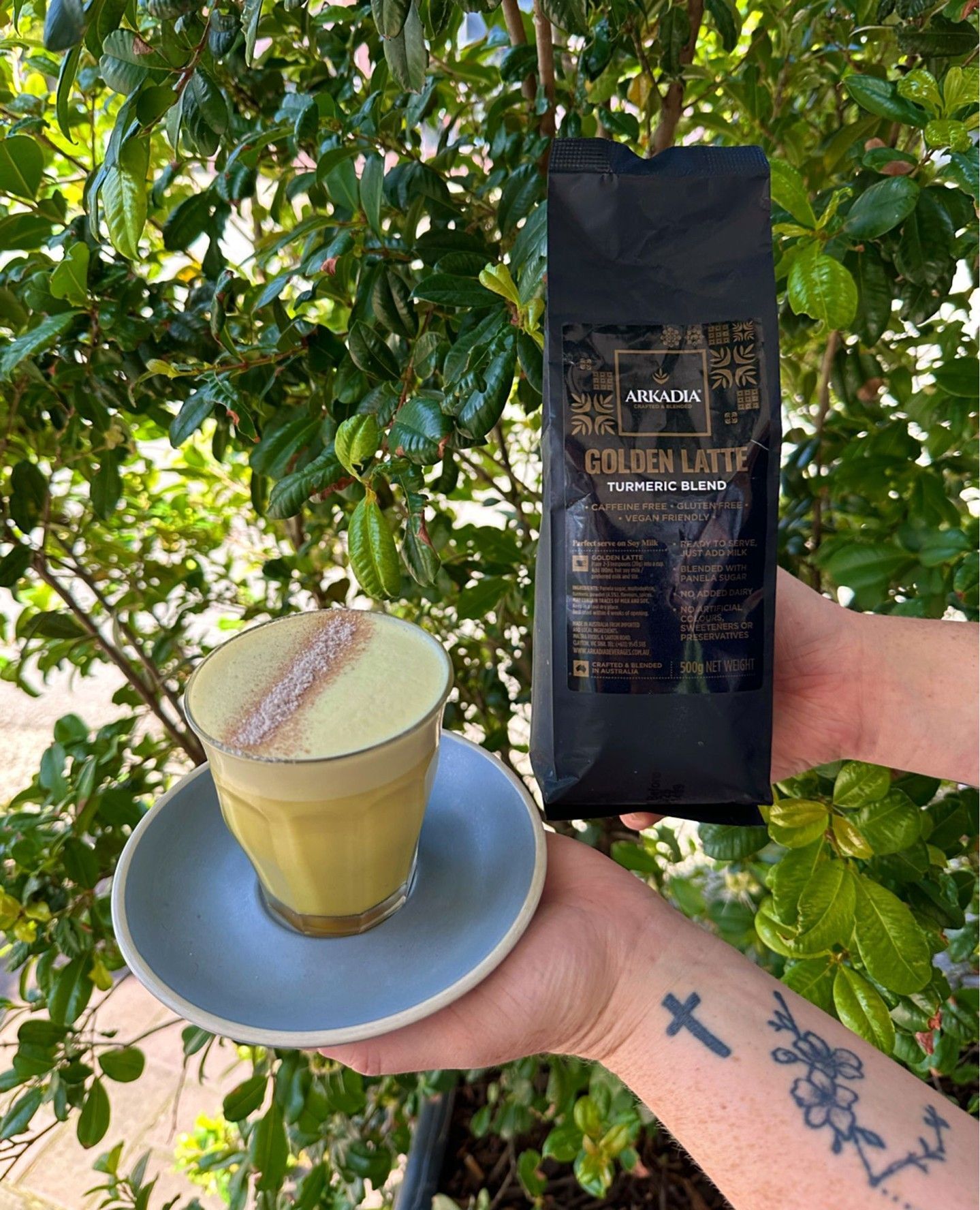 Person Holding a Golden Latte and a Black Coffee Bag in Front of Green Foliage — Drop In Espresso Bar in Wallsend, NSW