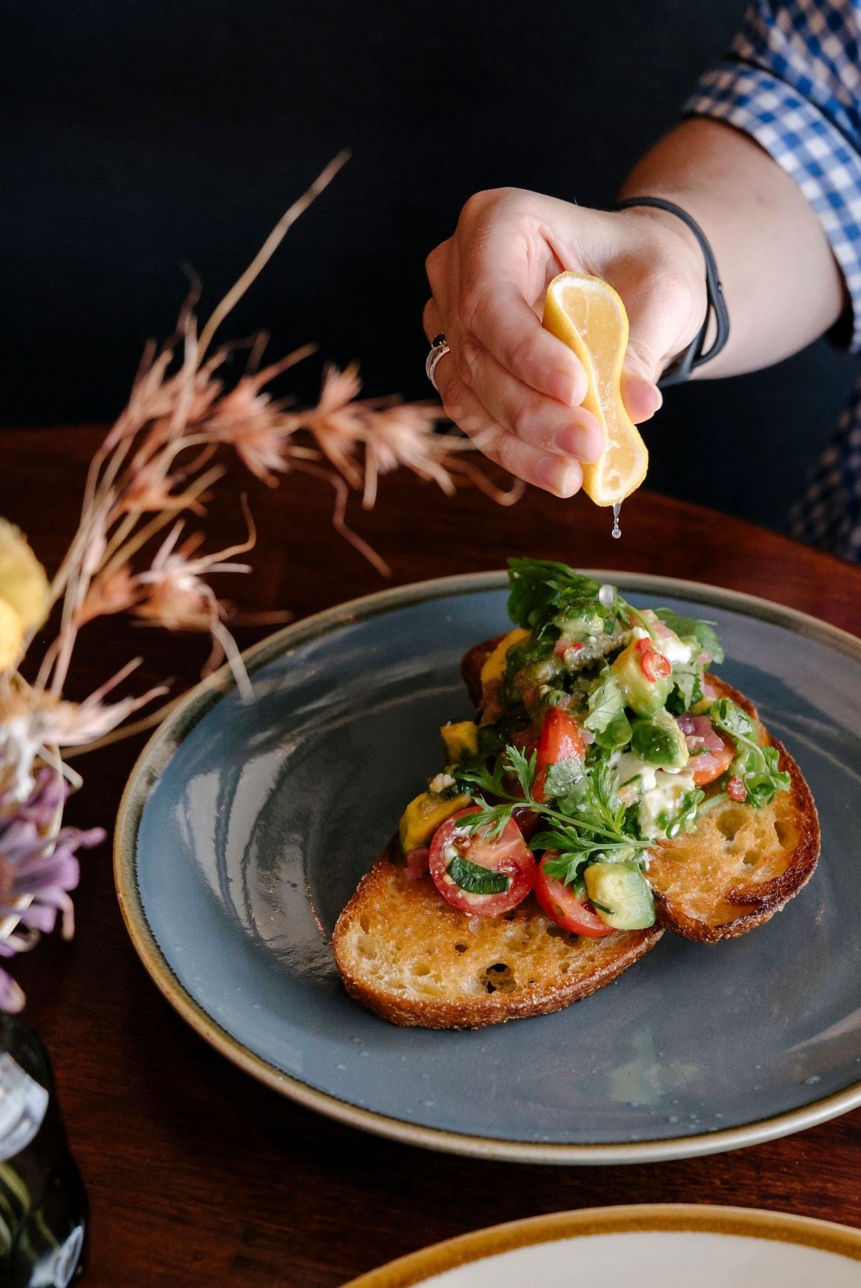 Hand squeezing lemon juice over a toast topped with tomatoes, herbs, and cucumber on a blue plate — Drop In Espresso Bar in Wallsend, NSW