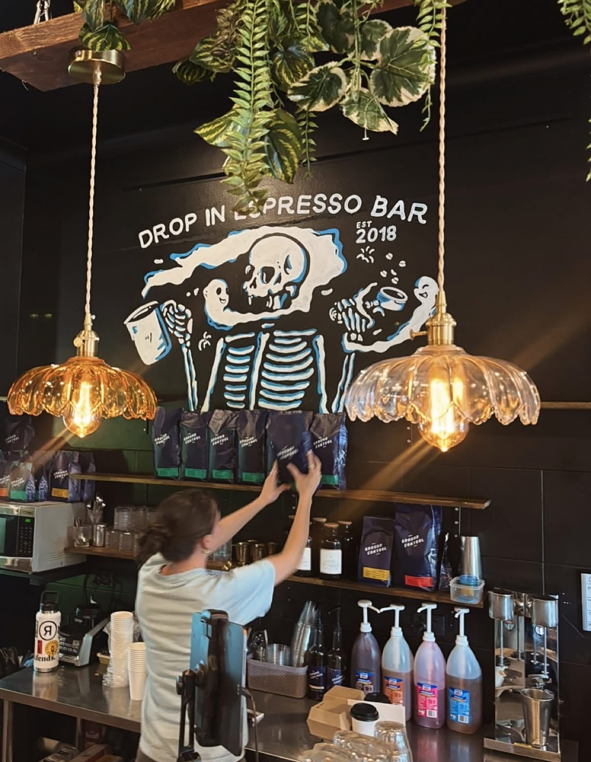 A barista reaching for coffee bags behind a counter in a coffee shop, with a skeleton mural on the black wall — Drop In Espresso Bar in Wallsend, NSW