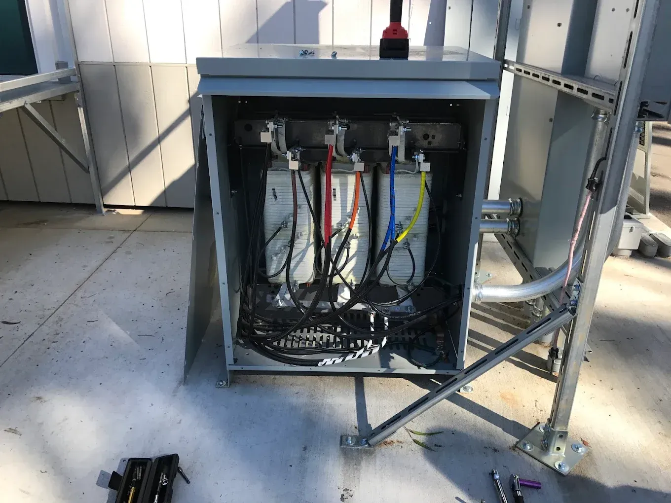 Open electrical box with three large cylinders, wires, and tools on the ground.