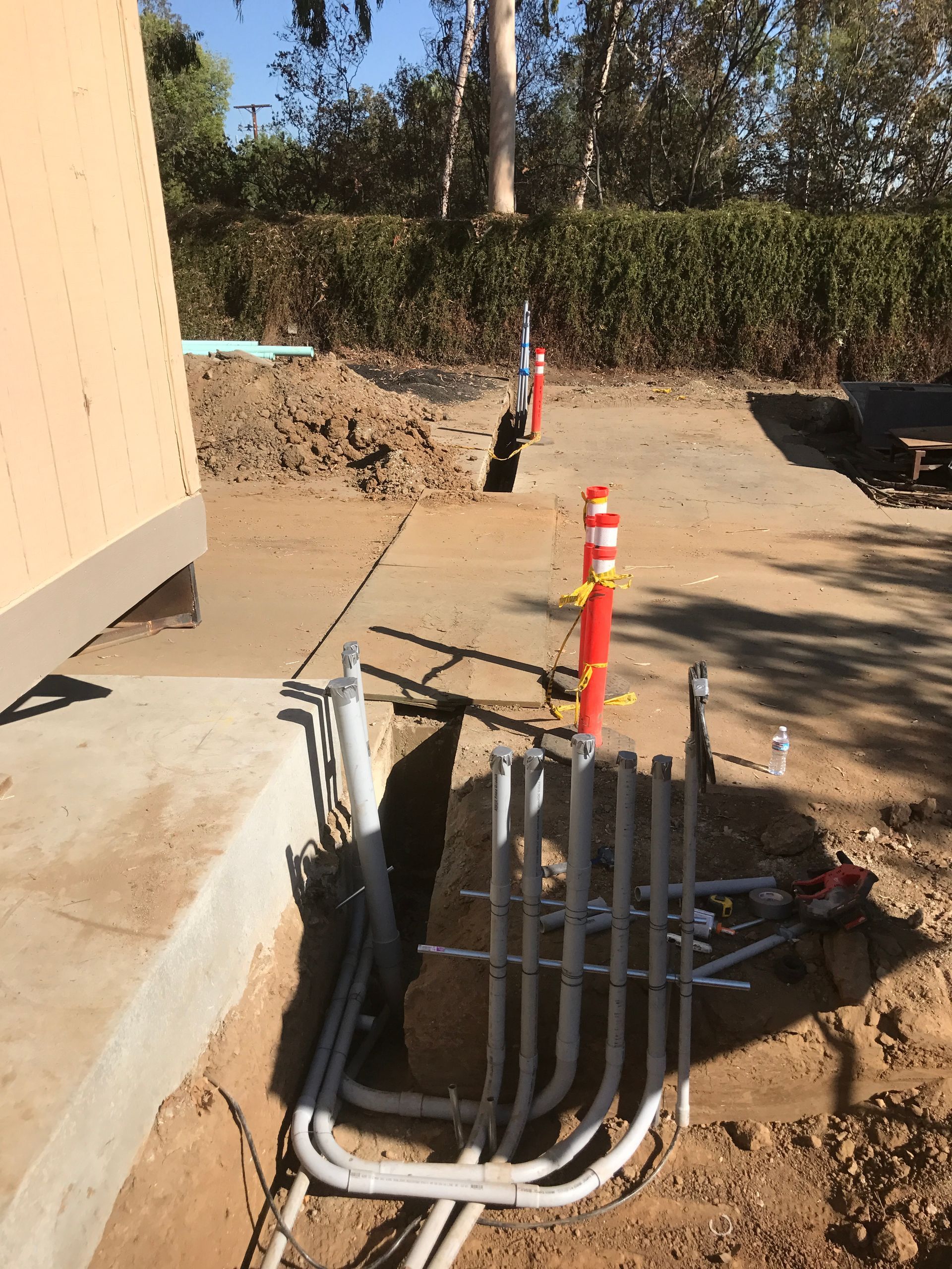 Electrical conduits exposed in a trench near a building. Red and white bollards mark the trench path.