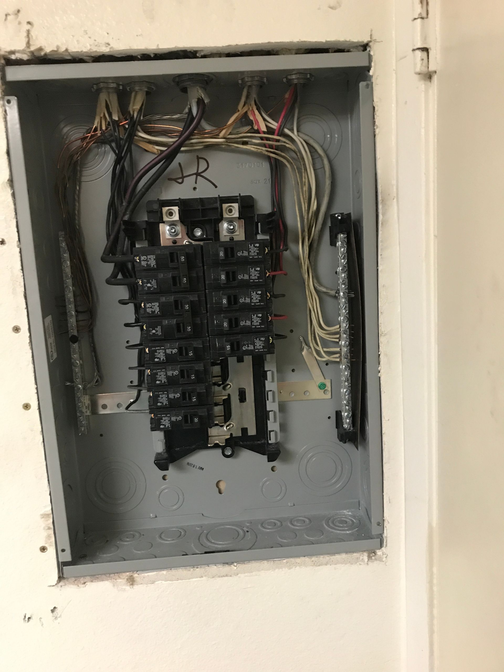 Electrical panel with exposed wires and circuit breakers, mounted in a wall.