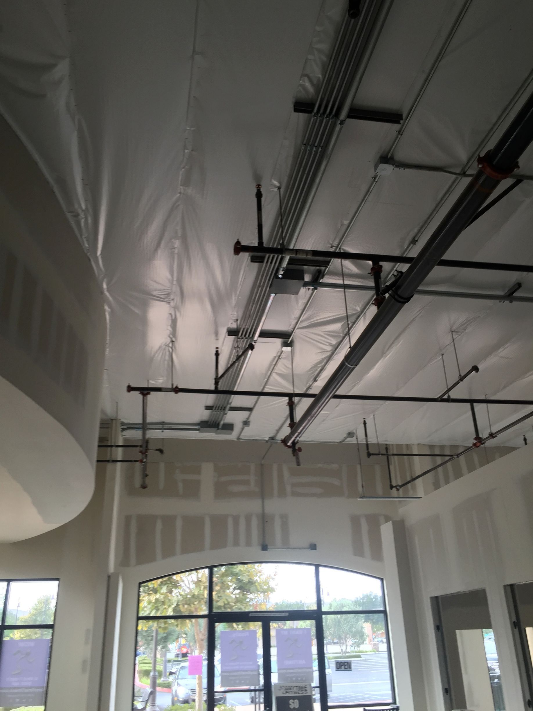 Interior view of a building under construction, showing unfinished ceiling with pipes, wires, and a large window.