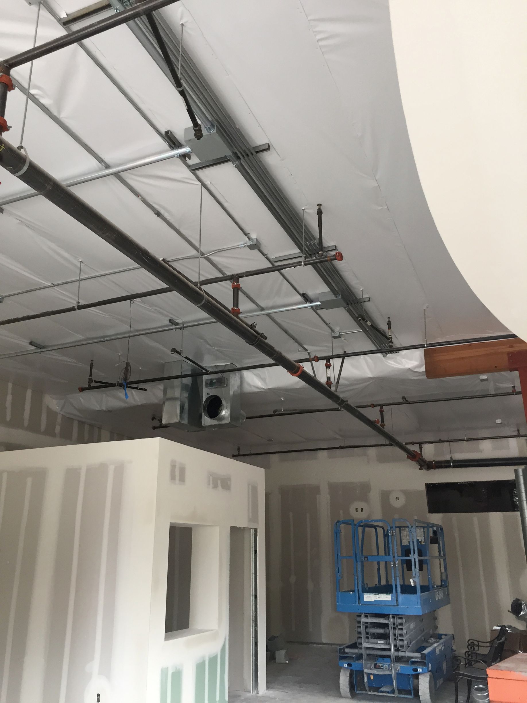 Interior construction site with exposed ceiling infrastructure.  A lift is present, and drywall is being installed.