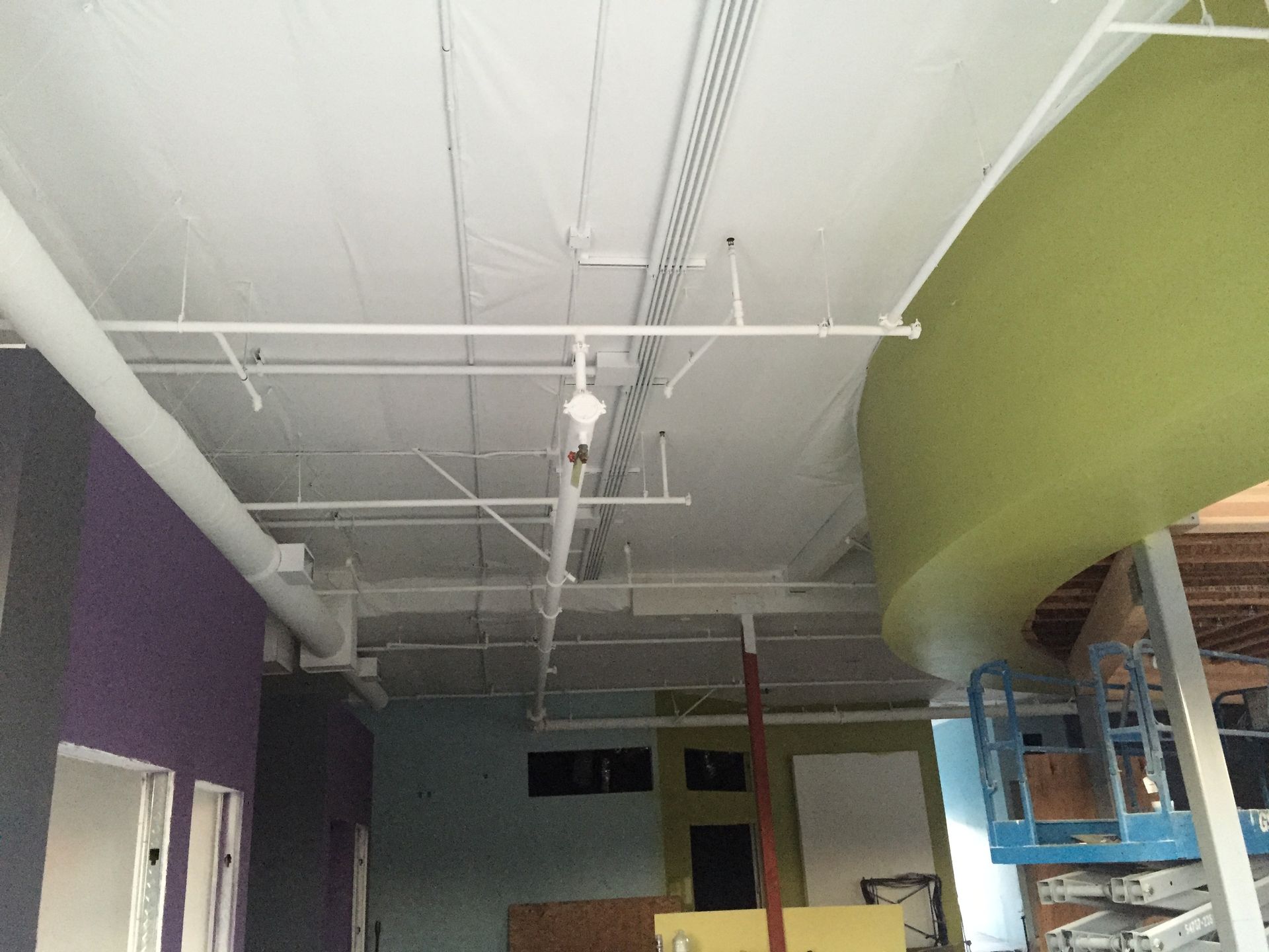 Construction site ceiling with white pipes, ductwork, and green wall.