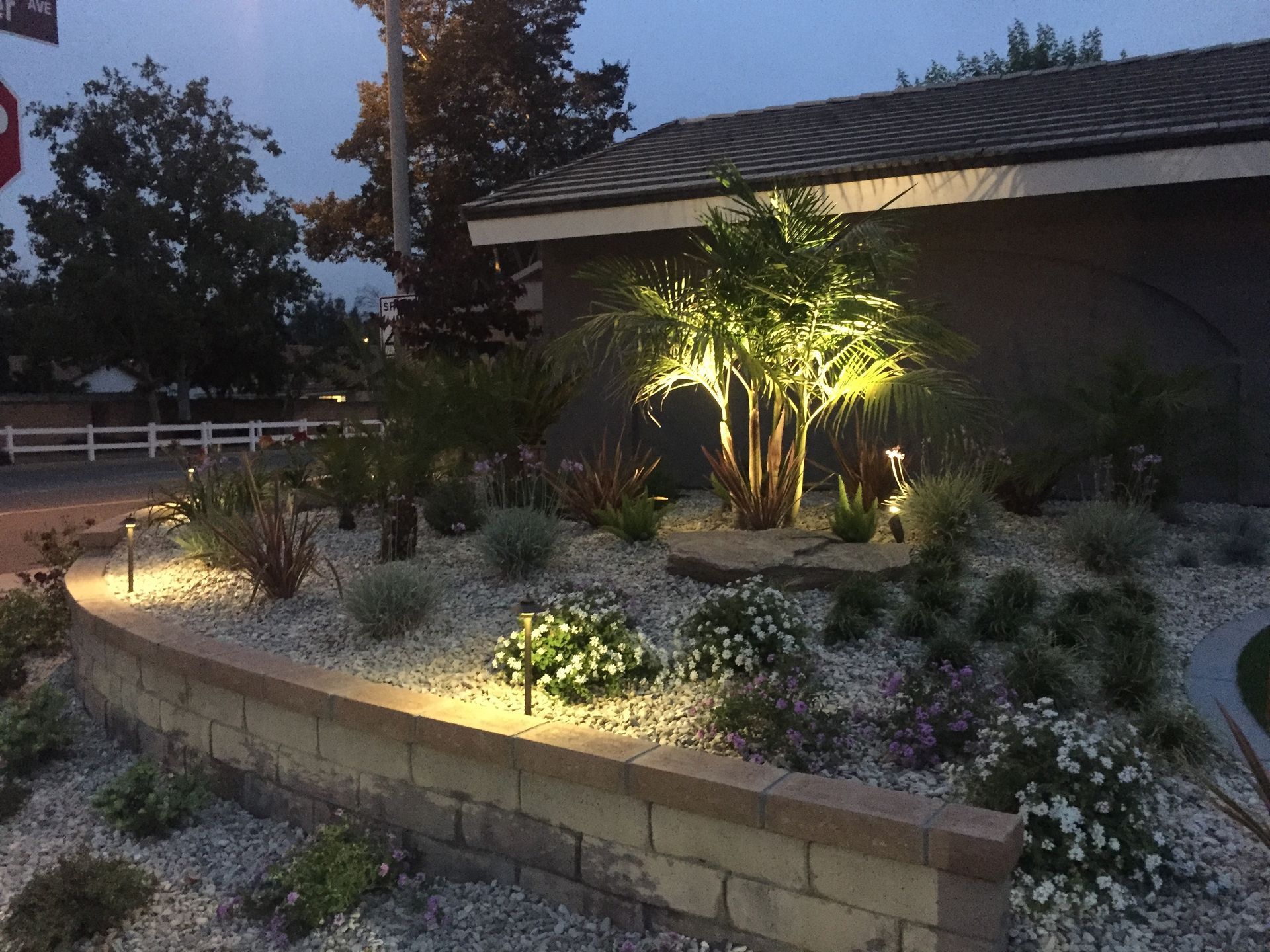 Night-lit landscaping with retaining wall, rock and plant beds. Tree lit from below. Building in background.
