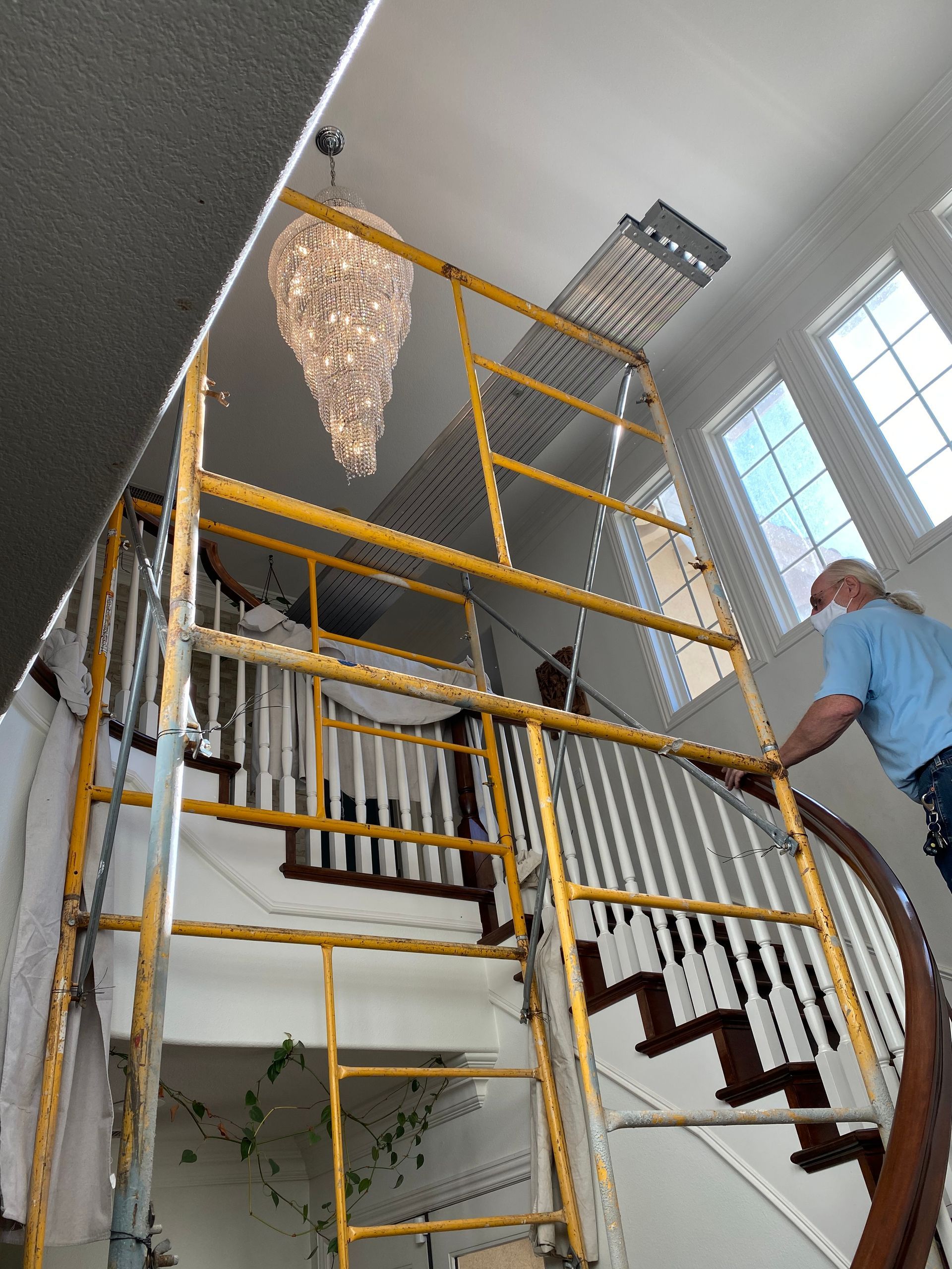 Yellow scaffolding erected near a winding staircase and chandelier. A person wears a mask and works on the scaffolding.