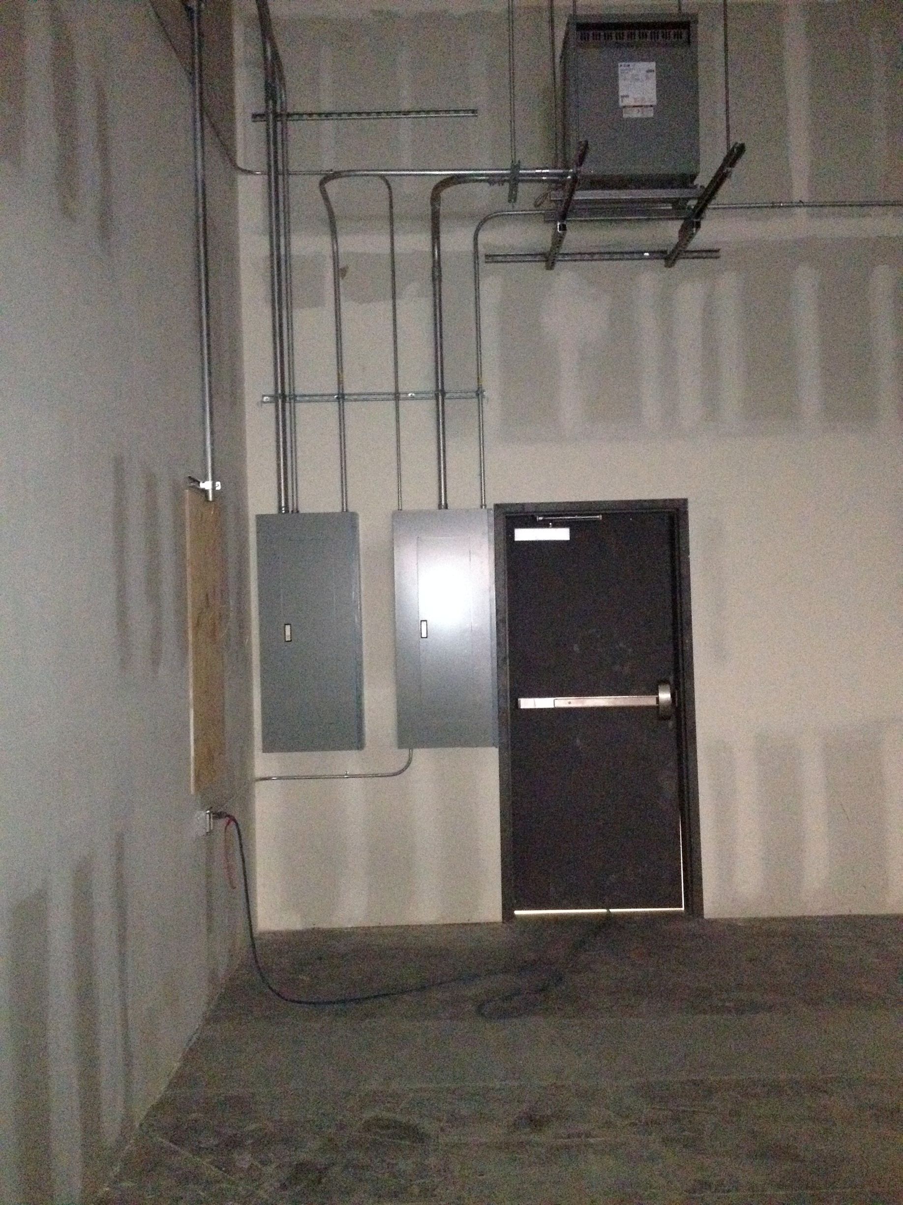 Electrical panels and a door in an unfinished room with conduit pipes.