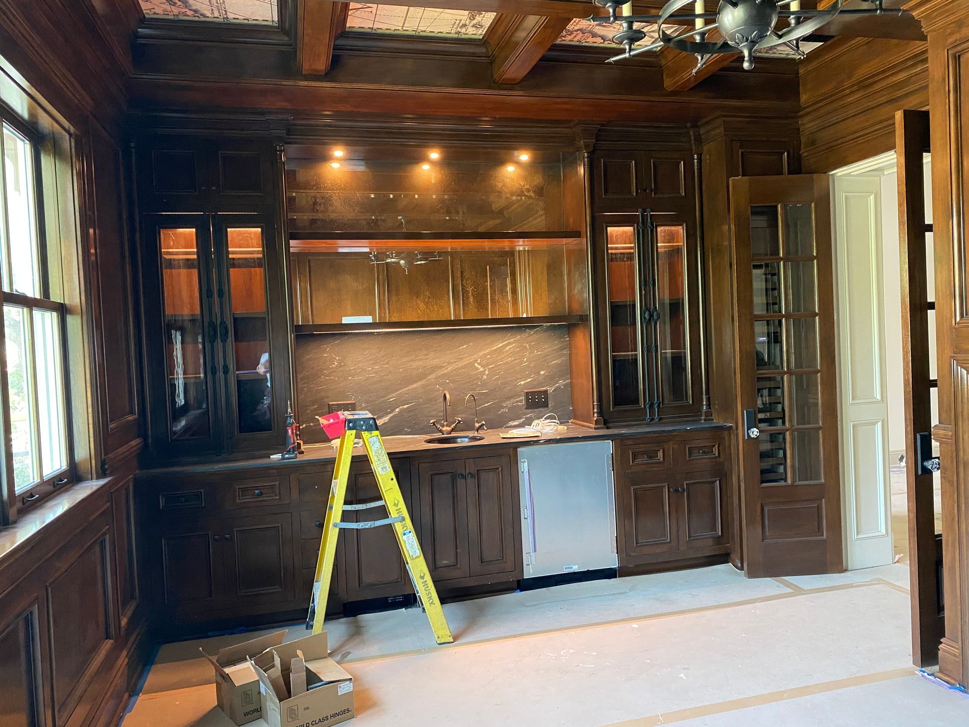 A dark wood home bar with cabinets, a sink, and a mini-fridge. A step ladder is in front of the counter.