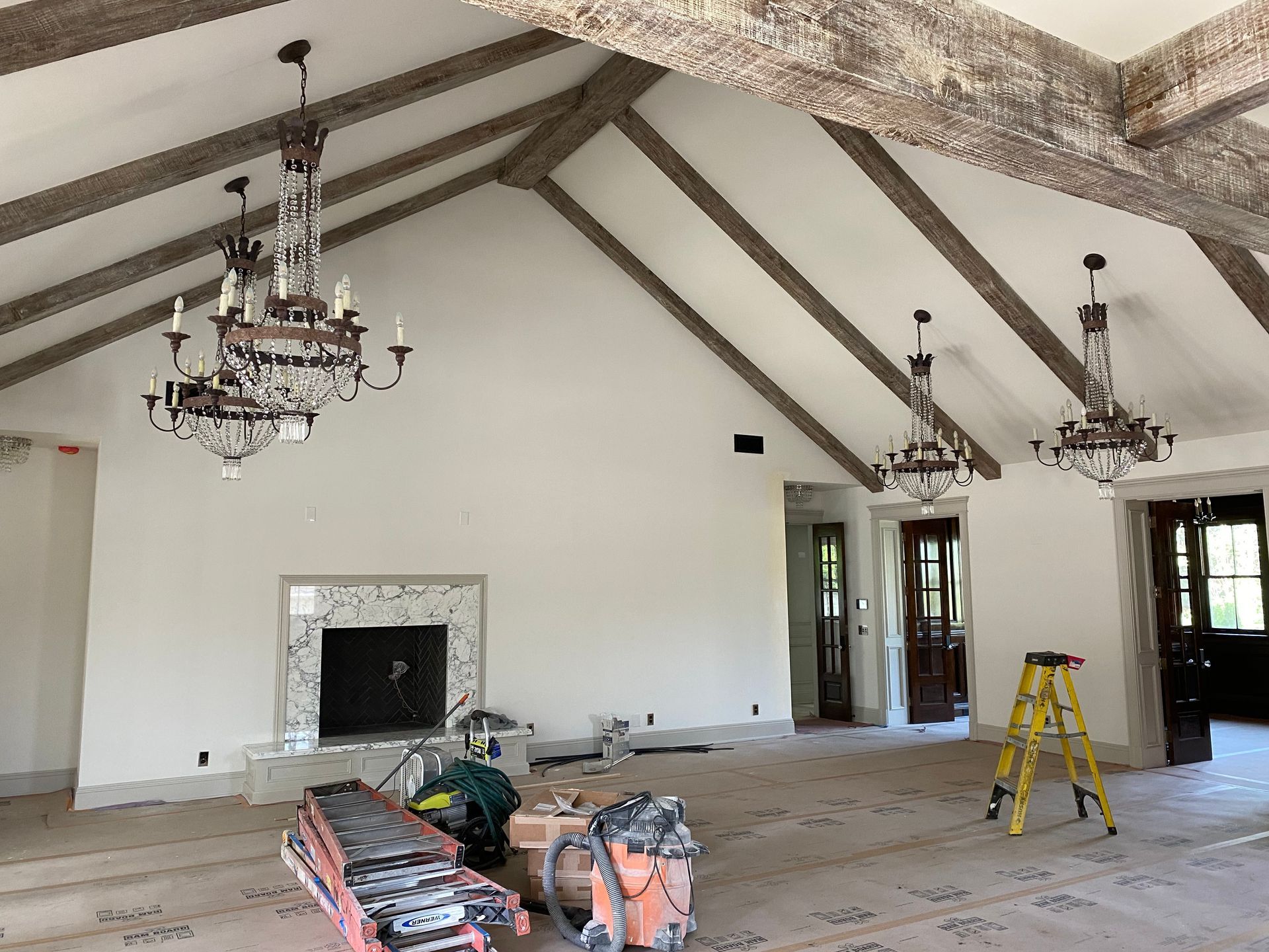 Empty room with high, angled ceiling, exposed beams, chandeliers, fireplace, and construction equipment.