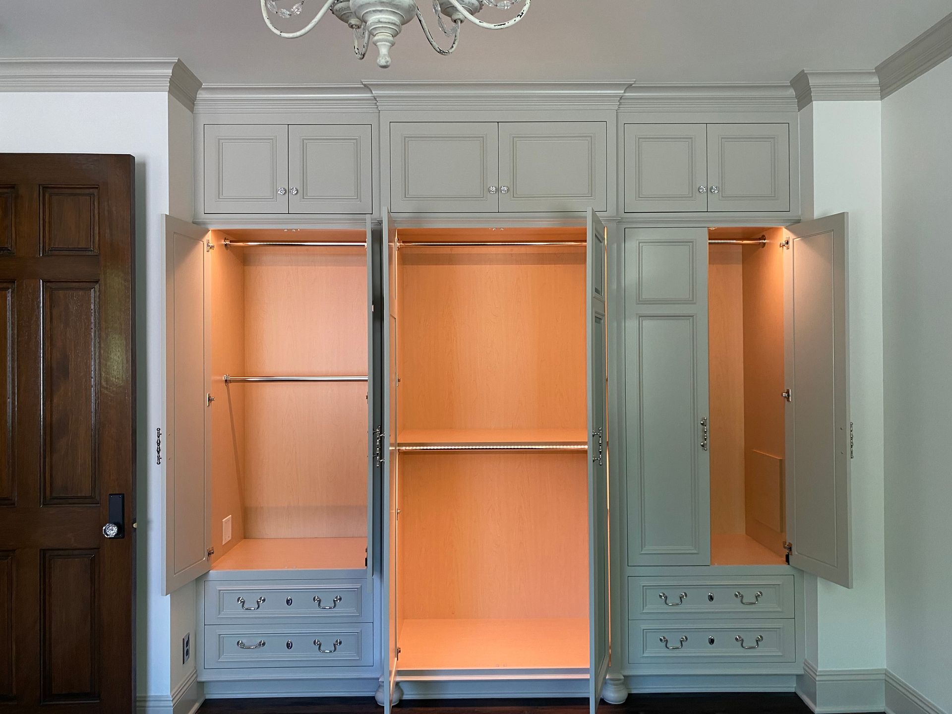 Built-in, grey wardrobe with open doors revealing clothes rods, shelves, and interior lighting. A dark wood door is to the left.