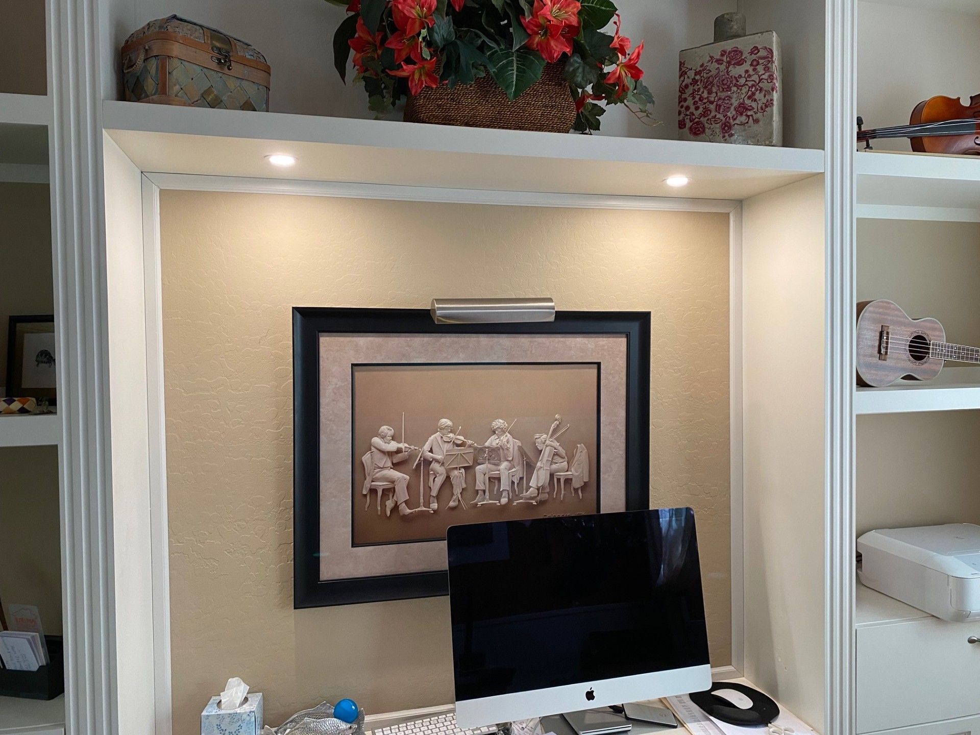 Built-in shelving with artwork, desk, and computer monitor. Tan walls, white shelves, and warm lighting.