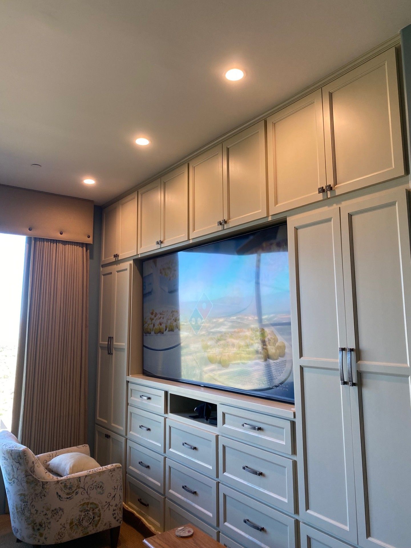 Built-in cream-colored cabinetry surrounds a large TV. Includes drawers, doors, and recessed lighting.