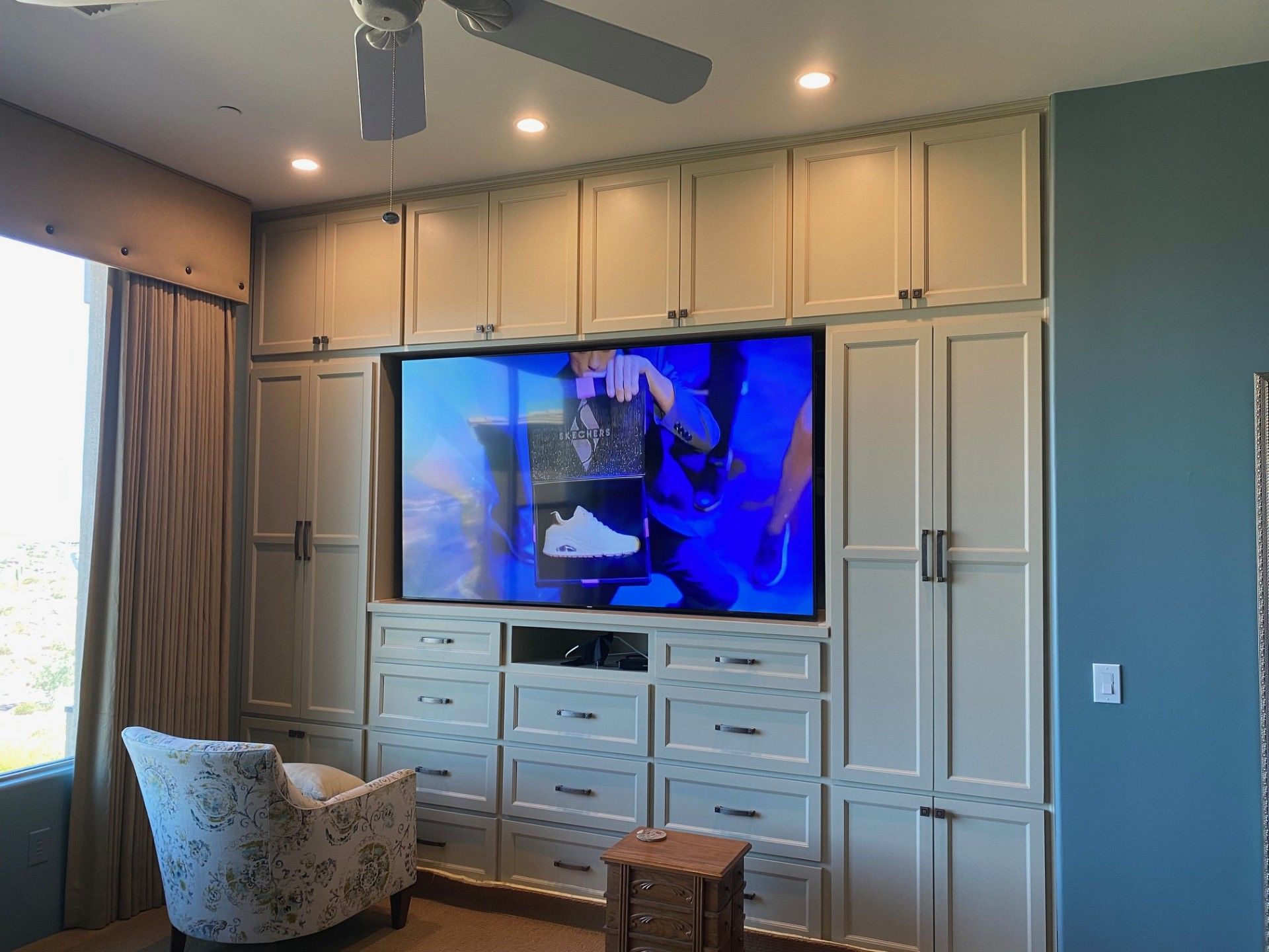 Built-in entertainment center with drawers, cabinets, and a large TV. A chair sits to the left, and there is a ceiling fan.