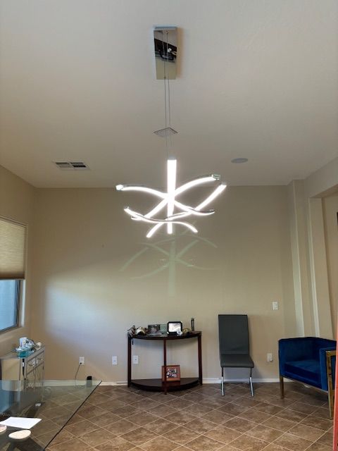 Modern chandelier over a table with electronics, a chair, and a blue velvet armchair. Beige walls, tile floor.
