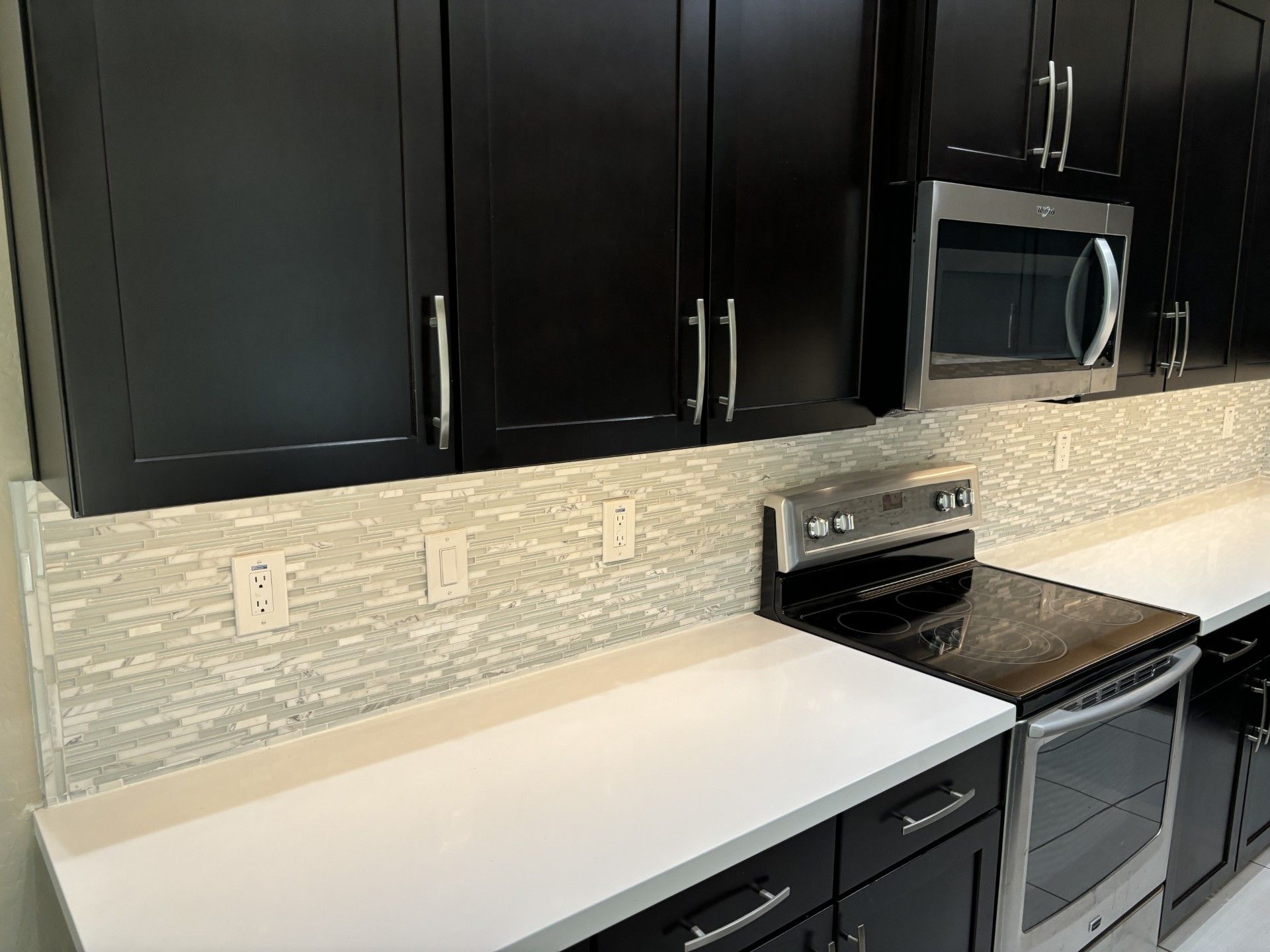 Black kitchen cabinets, stainless steel appliances, and white countertops. Light-colored backsplash.
