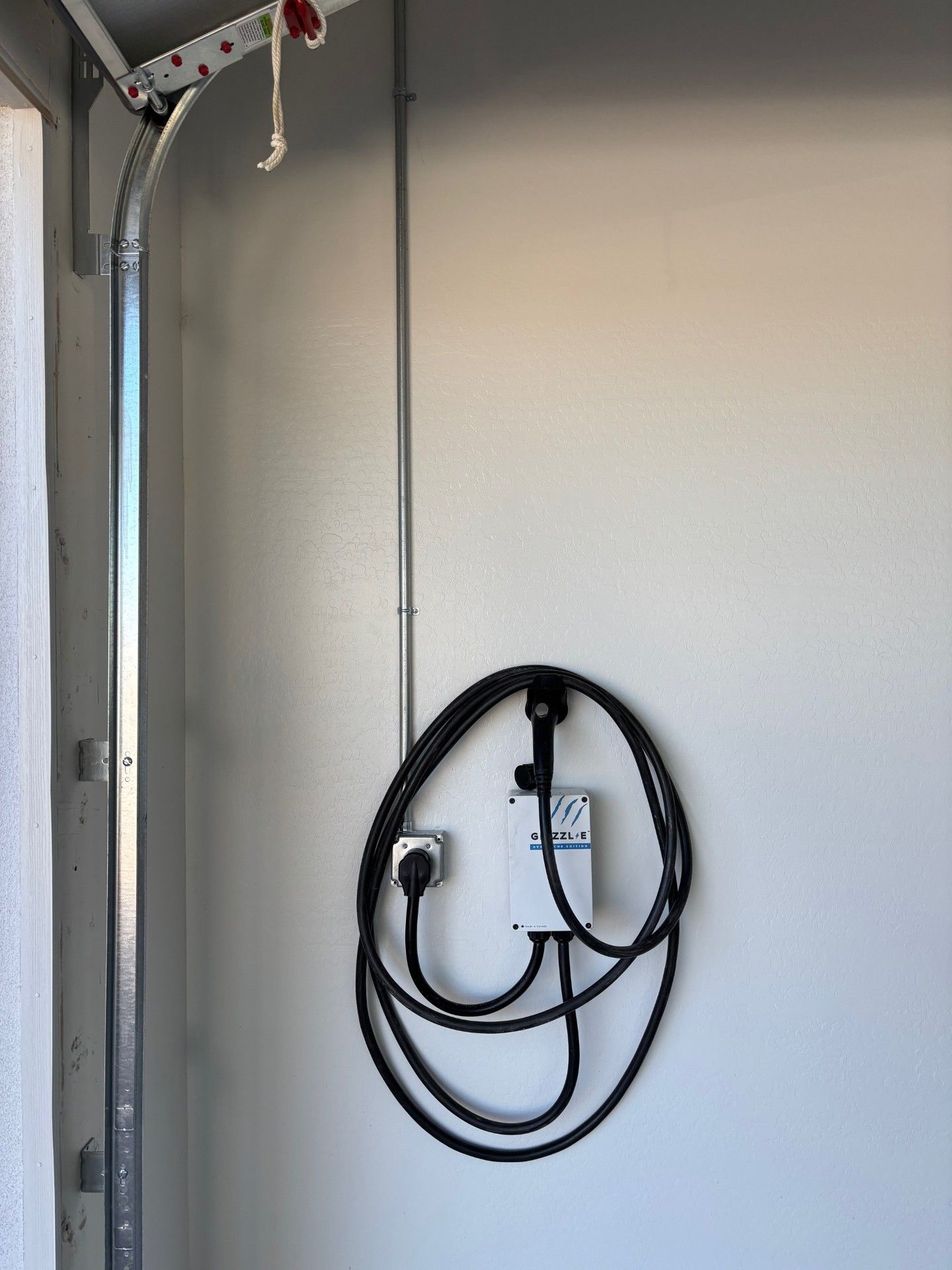 An electric vehicle charger mounted on a white wall in a garage. Black cable loops down to a wall outlet.