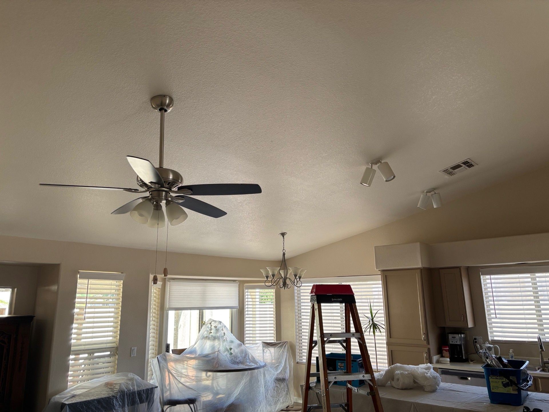 A room with a textured ceiling, a ceiling fan, a ladder, and a covered table.