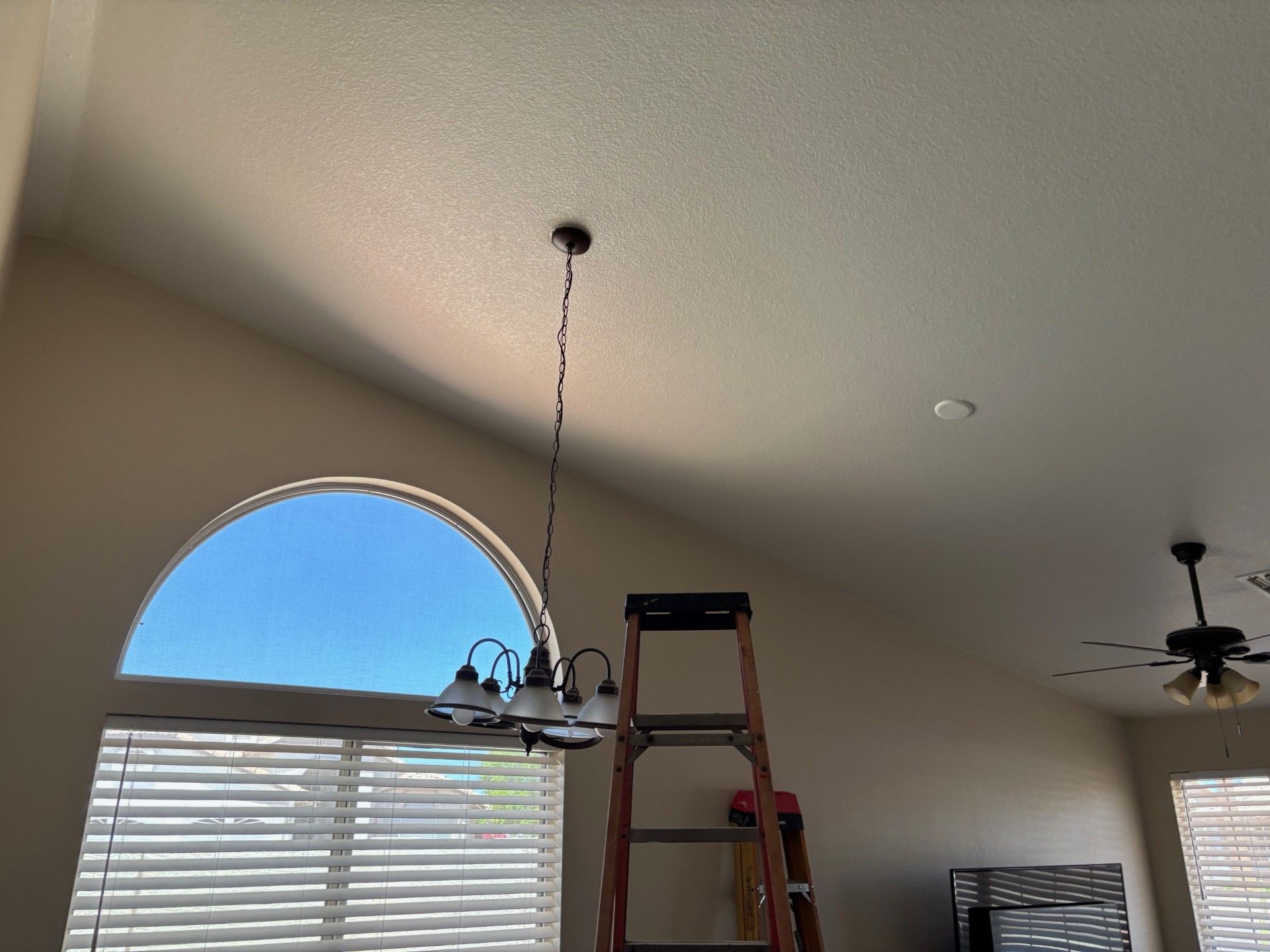 Ladder below a chandelier against a high, sloped ceiling with a large arched window and a ceiling fan.