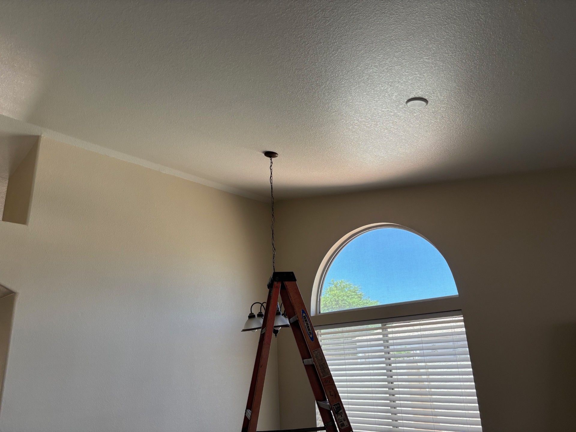 Ladder beneath a light fixture on a beige ceiling, arched window with a view of sky and trees.
