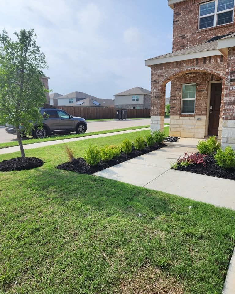 A suburban brick home with a concrete walkway, landscaped garden beds, a young tree, and a parked SUV on a sunny day.
