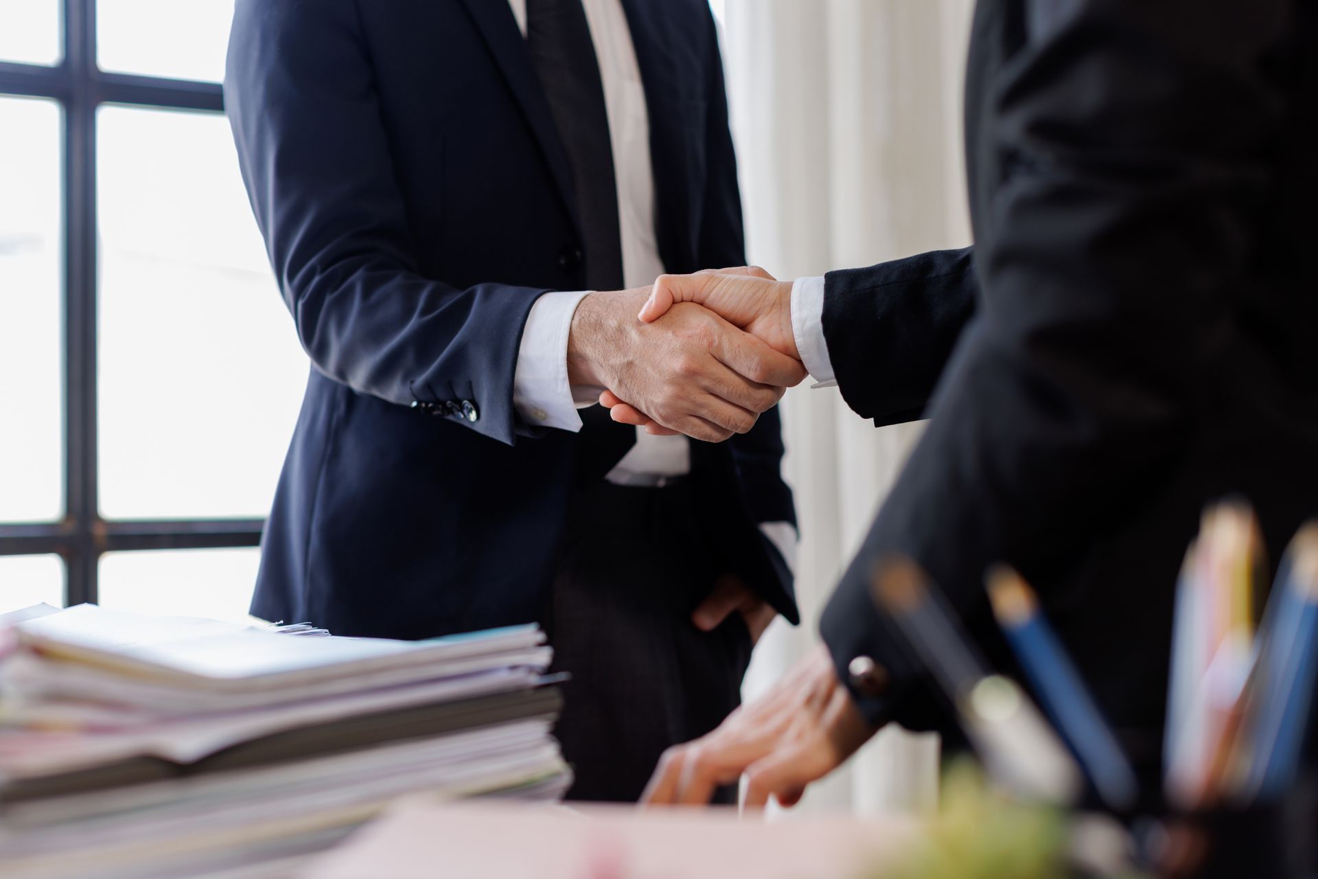 Two people in suits shaking hands at a desk, near a window and documents.