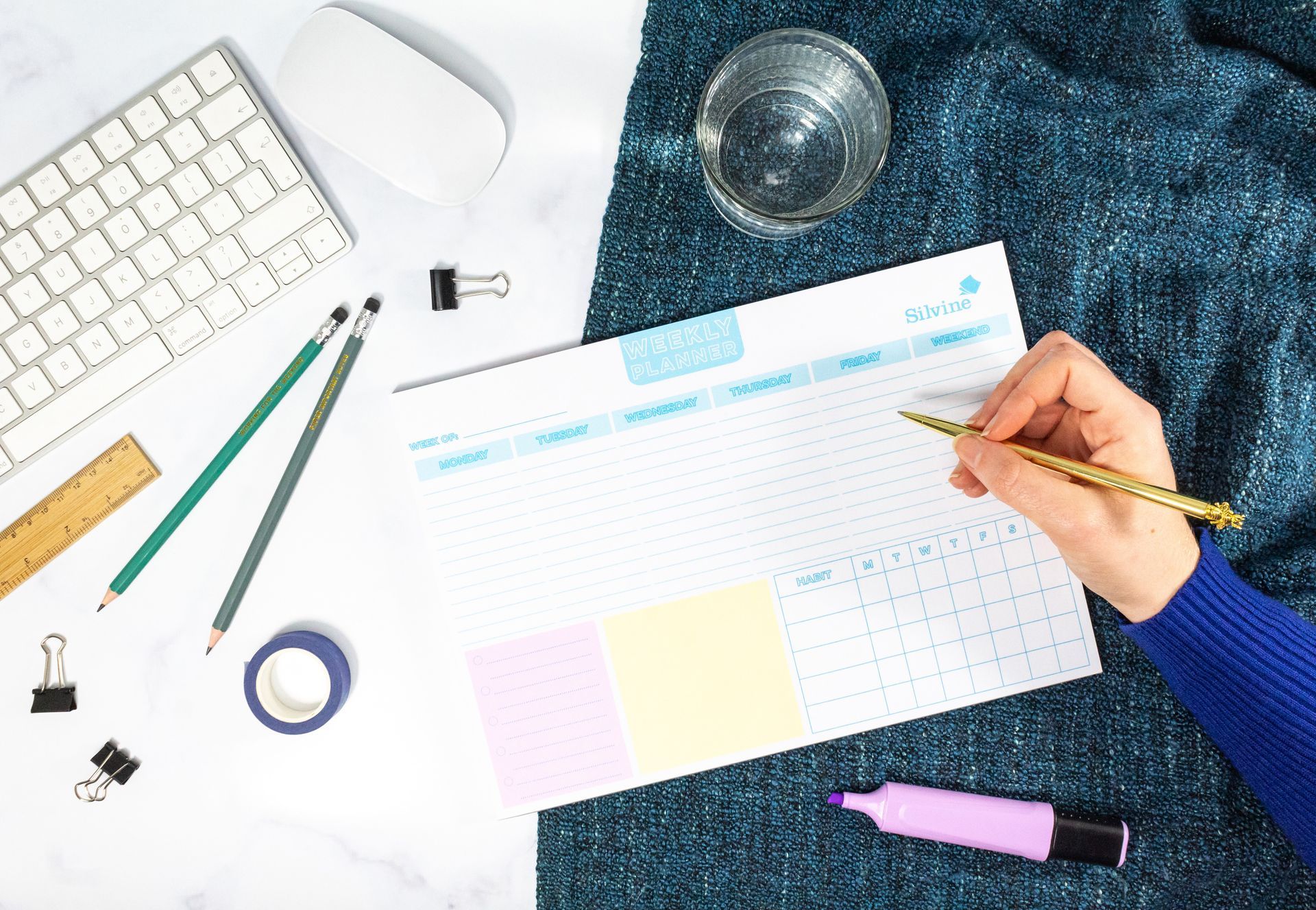 A weekly planner pad on a desk with keyboard, stationery items, water and hand about to write on the pad.